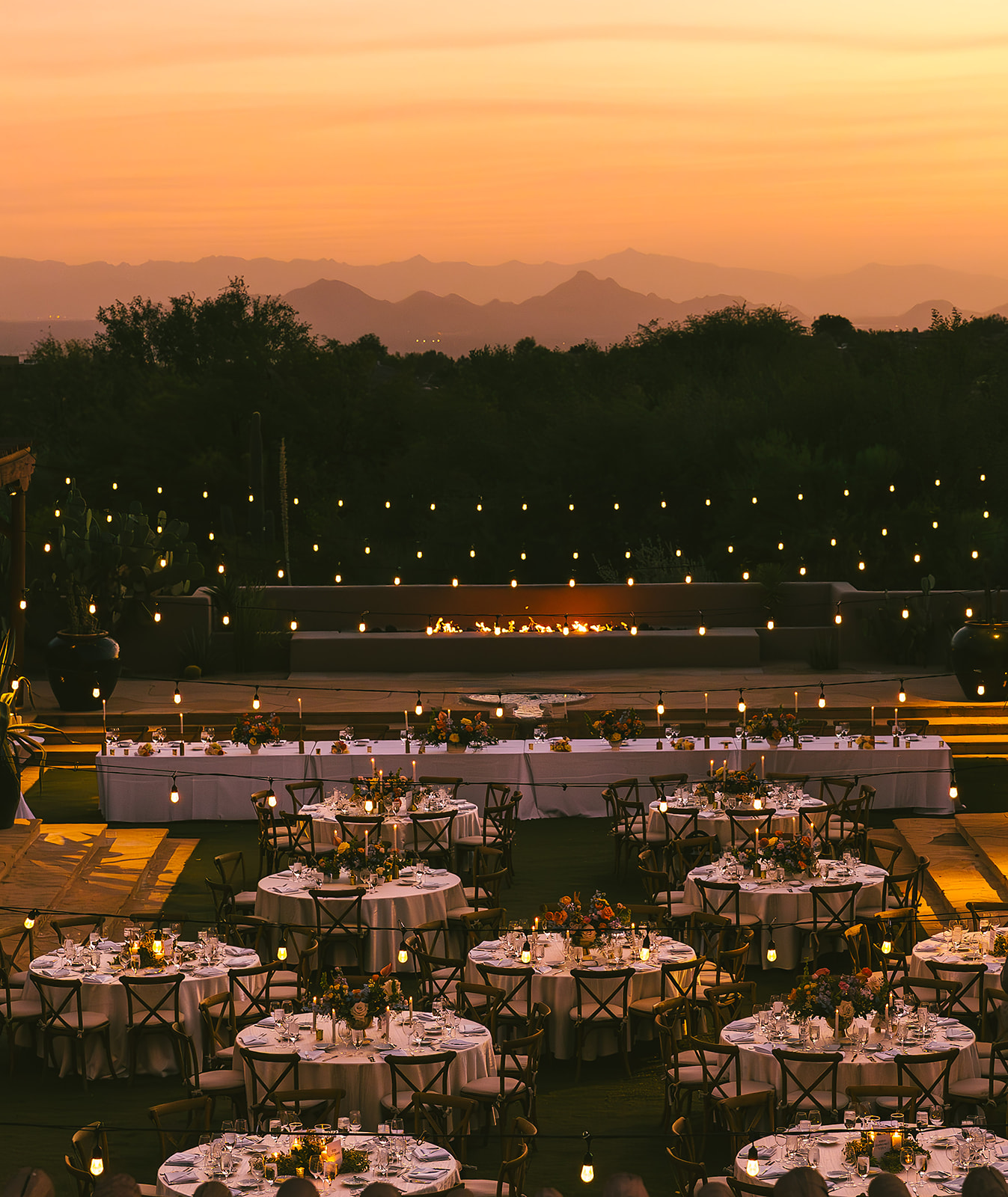 Outdoor dining setup with tables and chairs, sunset sky with mountains in the background, string lights overhead, and a scenic view.