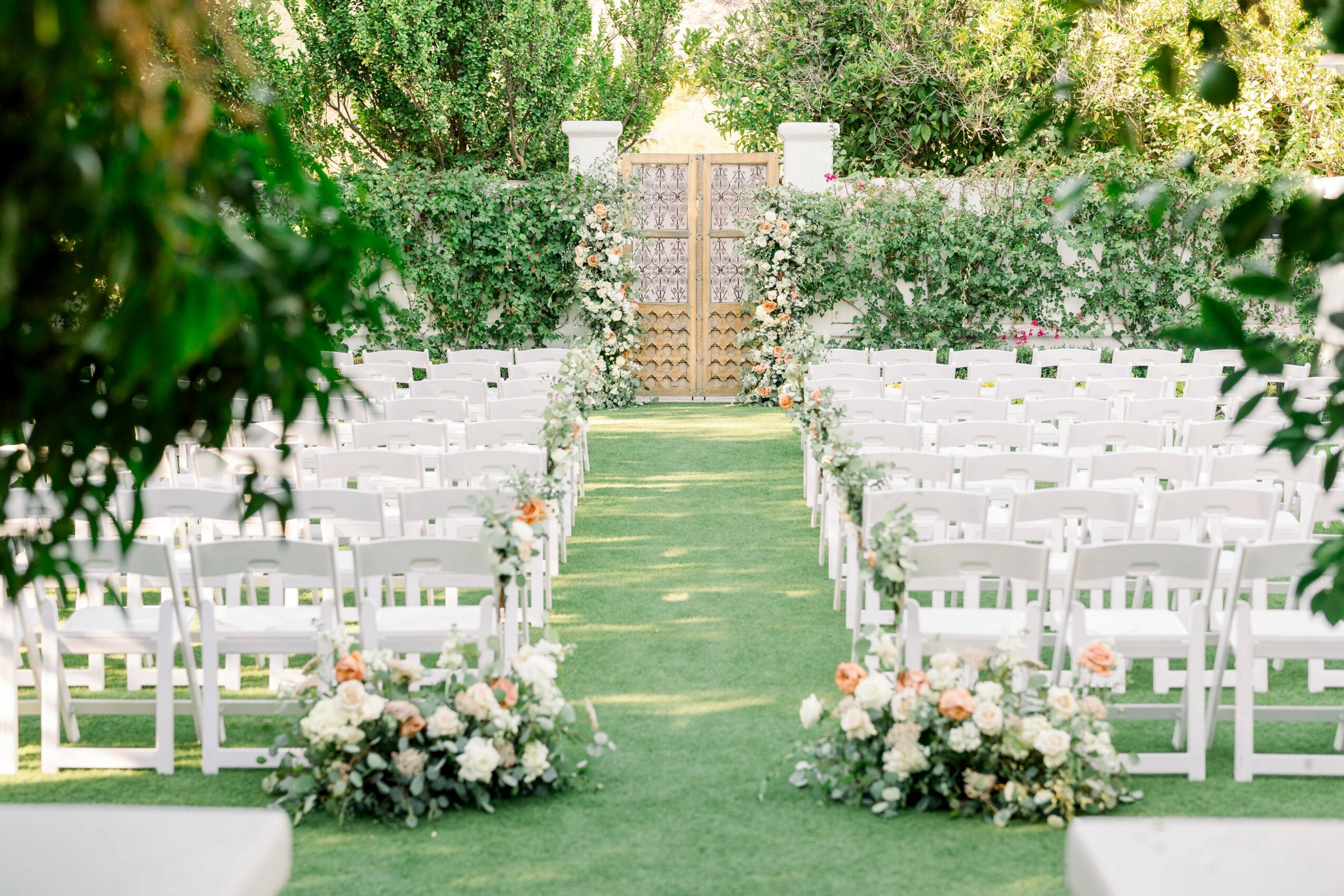 Outdoor wedding ceremony setup with white chairs, floral arrangements, and a green aisle leading to a wooden gate, surrounded by trees.