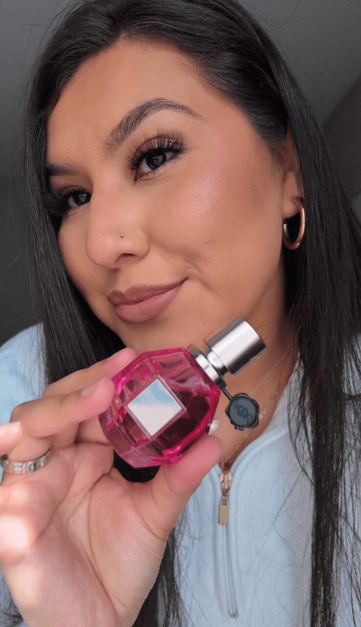 Woman with dark hair and hoop earrings holding a pink perfume bottle, smiling slightly, in a close-up portrait.
