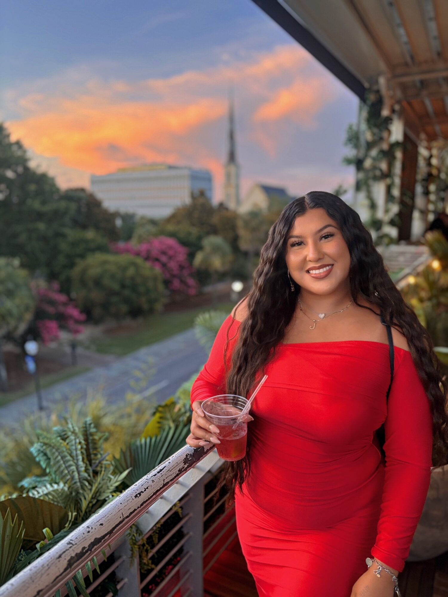 Woman in red dress smiling on balcony with cityscape and sunset in background