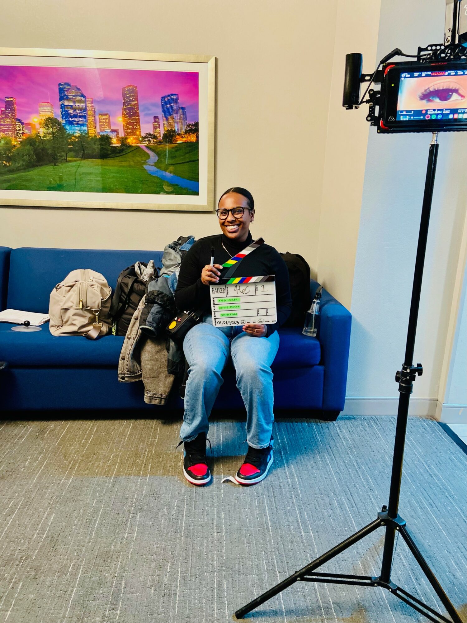 Person sitting on a blue couch holding a clapperboard, smiling, with a colorful cityscape picture on the wall behind.