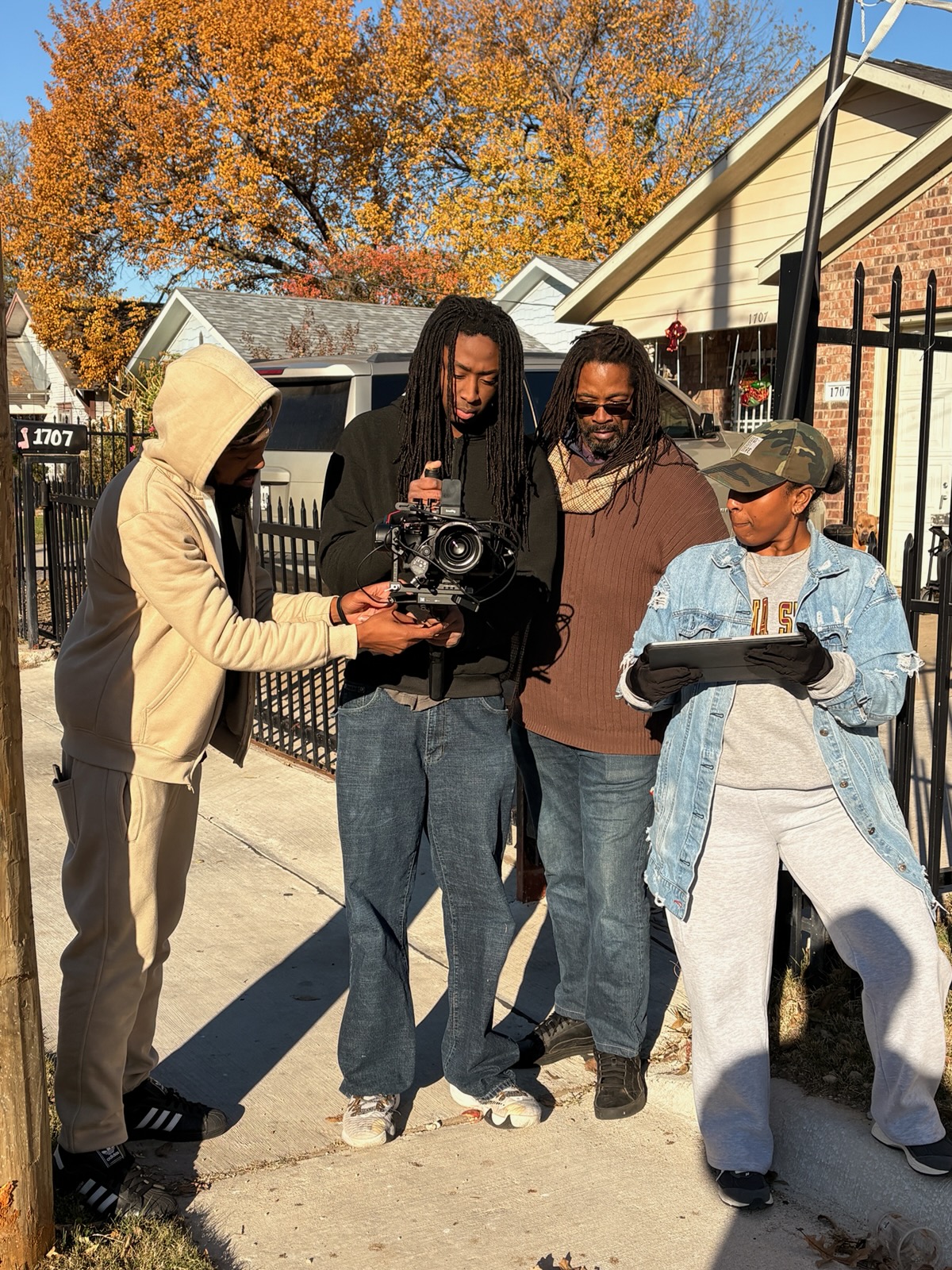 Four people stand on a sidewalk in front of houses, one holding a camera, others looking at a tablet, autumn trees in background.