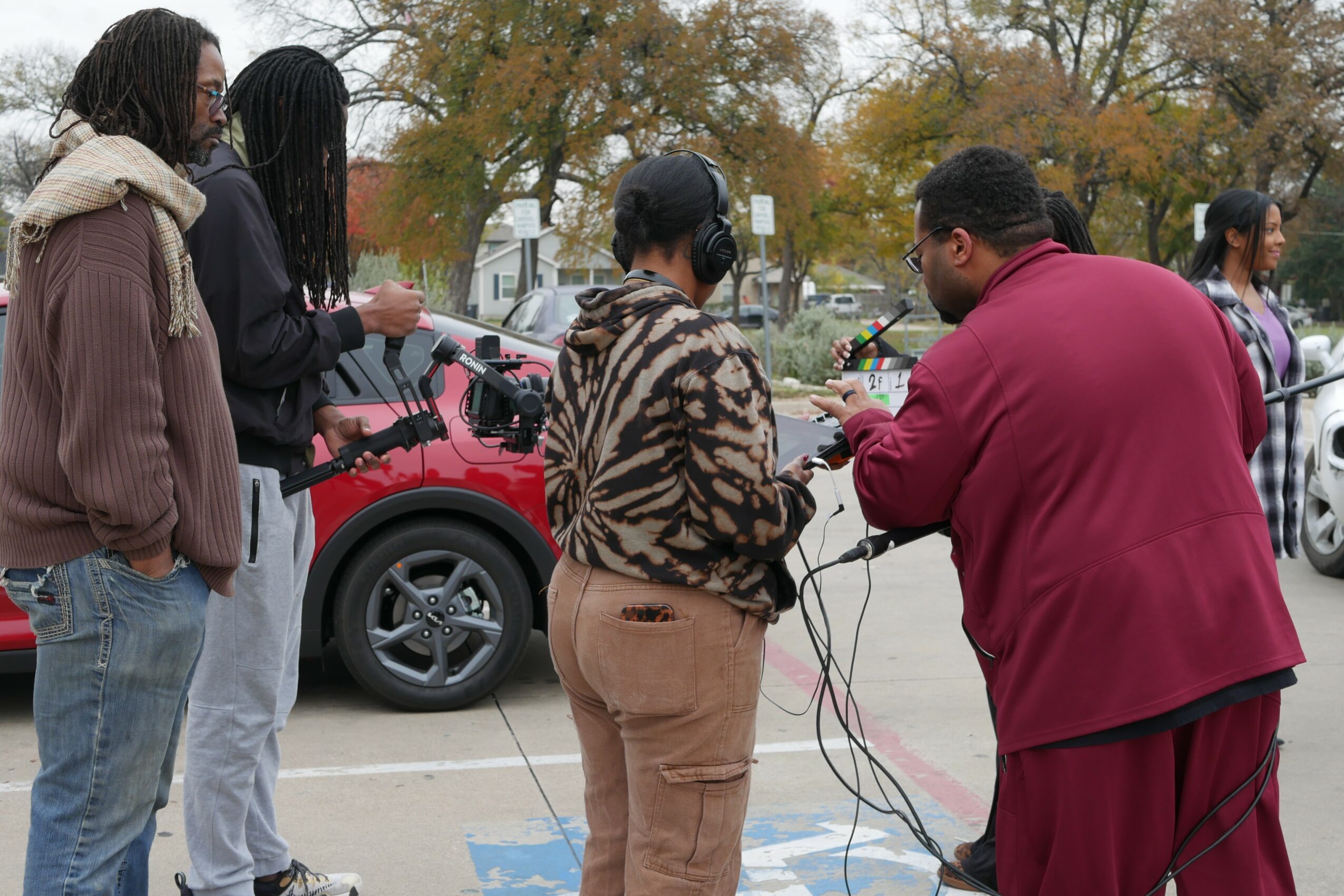Group of people standing outdoors near parked cars, some holding microphones and wires, with trees in the background.