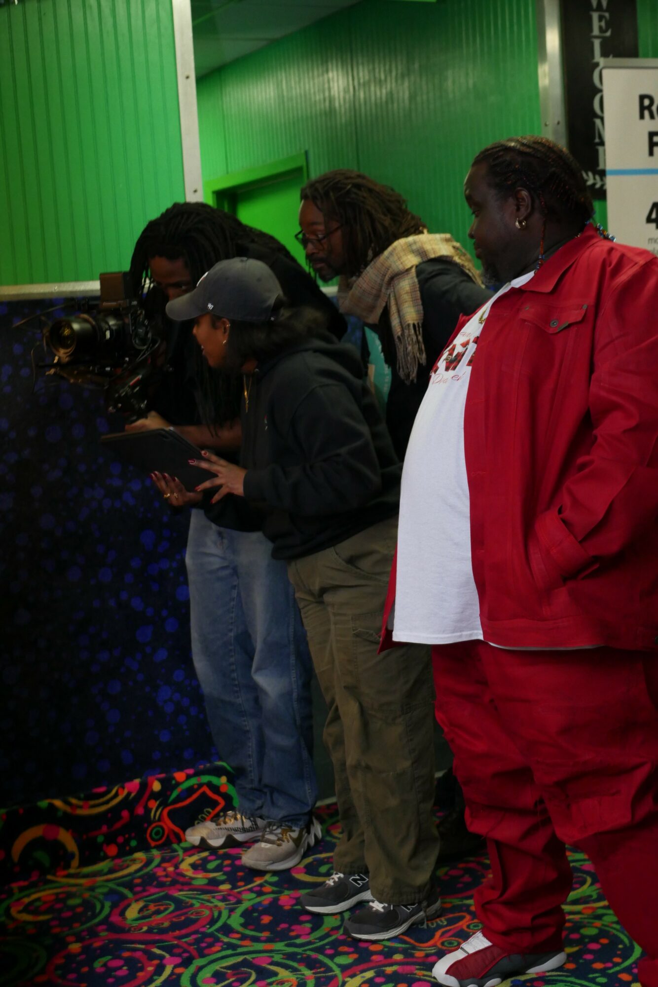 Group of people standing indoors near a green wall, some looking at a device, colorful patterned carpet on the floor.