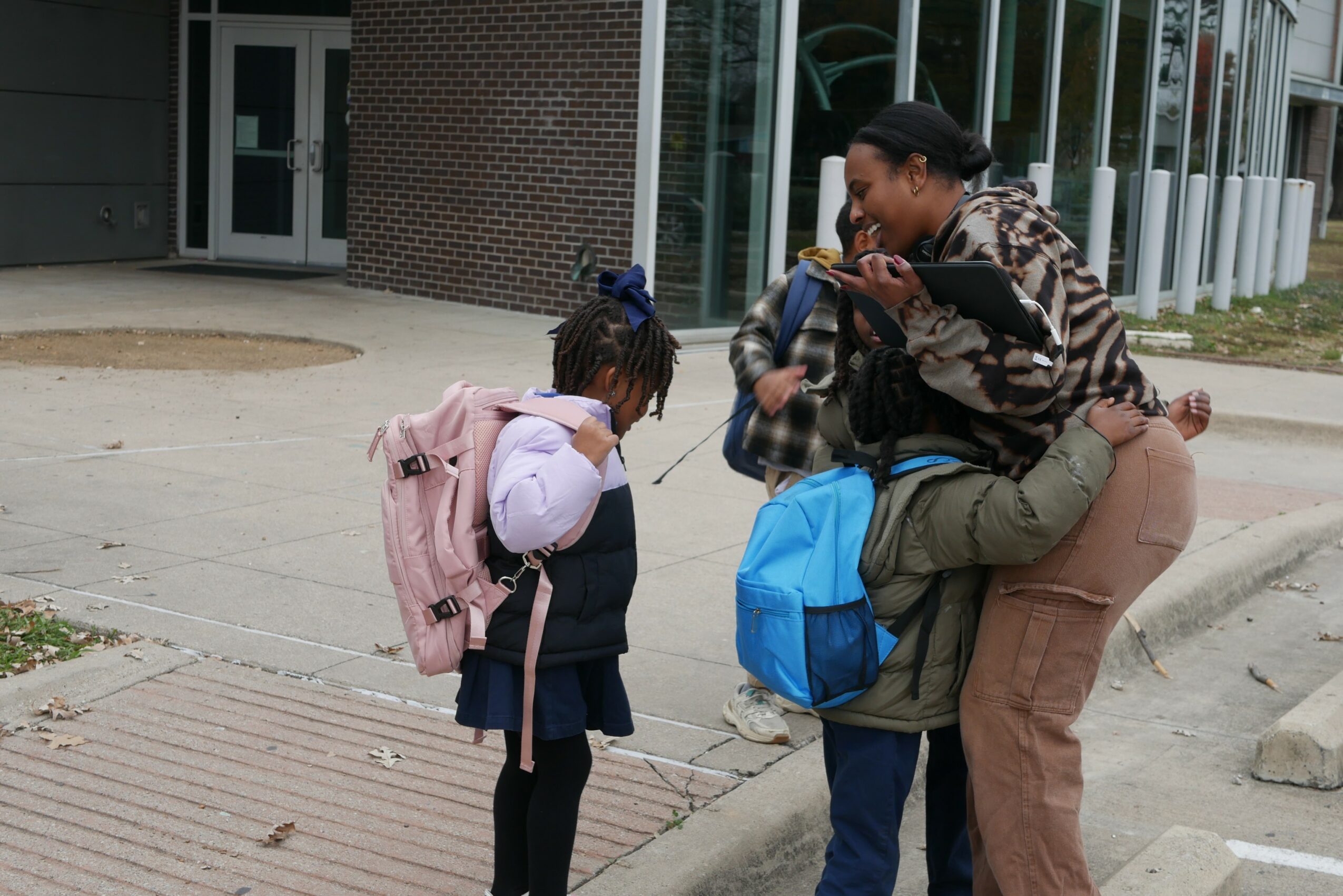 Adult woman crouching and talking to two children with backpacks outside a building.