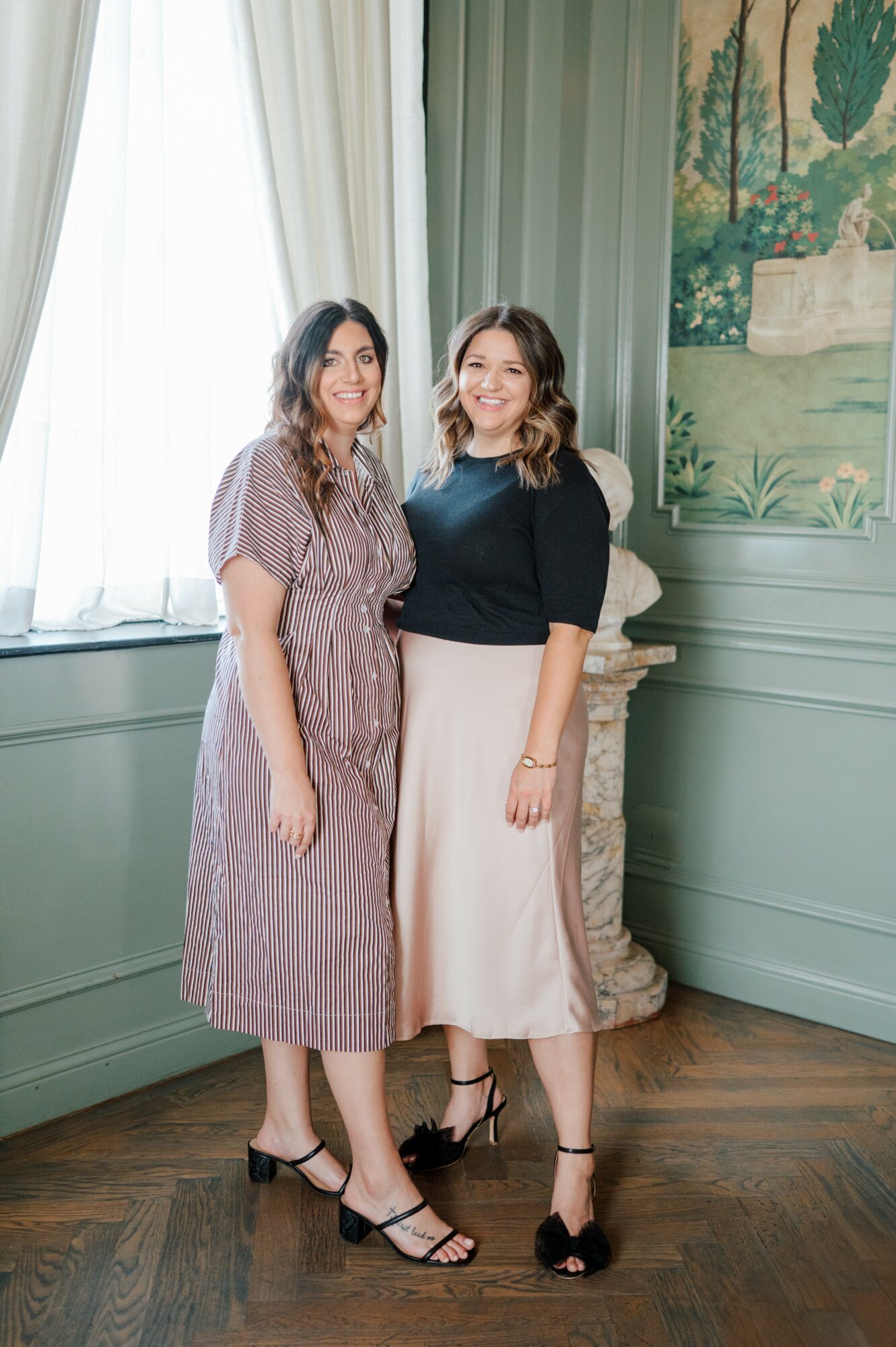 Two women standing indoors near a window with green walls and a decorative plant, smiling at the camera.
