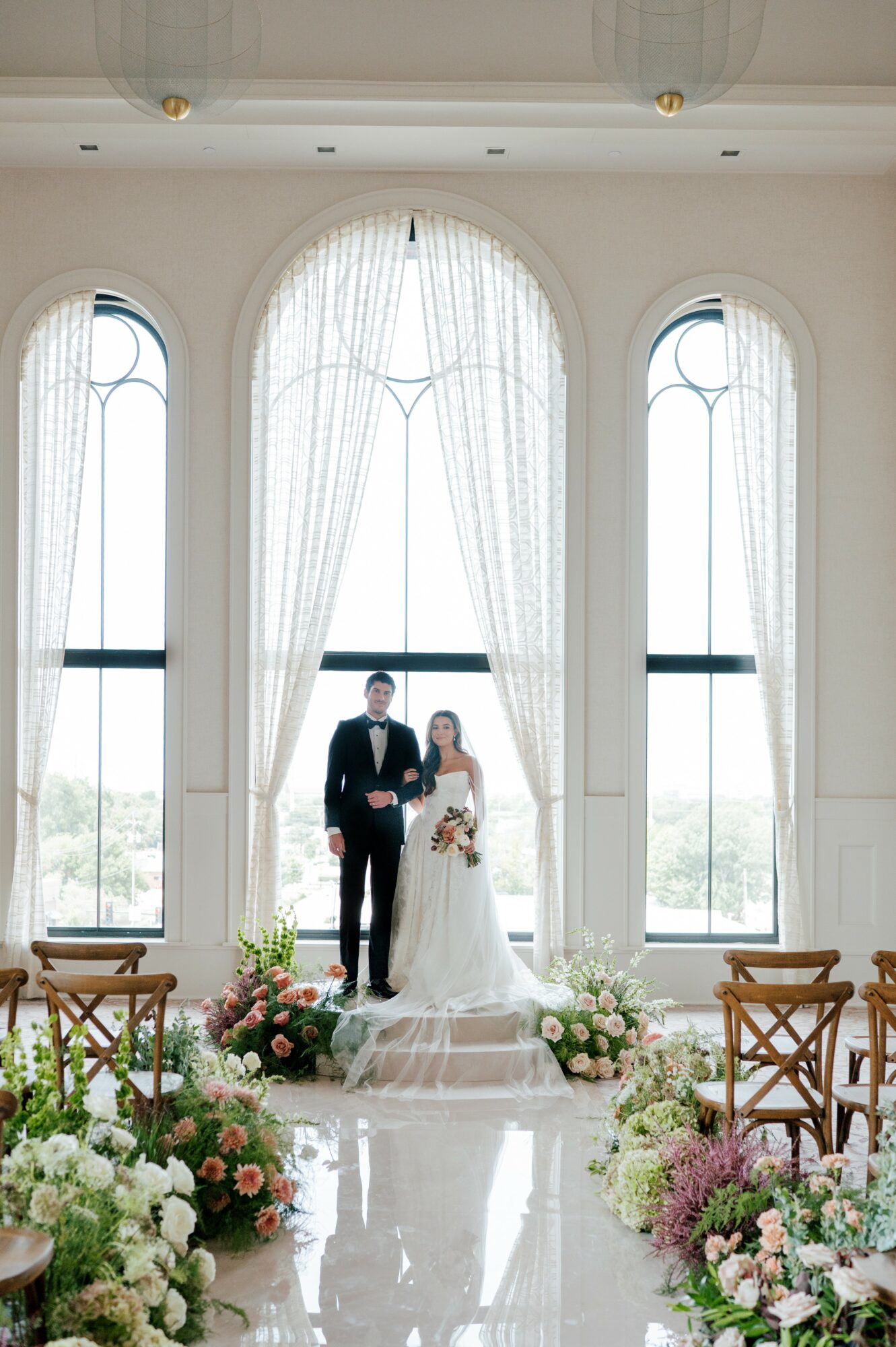 Bride and groom standing on a decorated platform in front of large arched windows with white curtains, floral arrangements on the floor, and wooden chairs on sides.