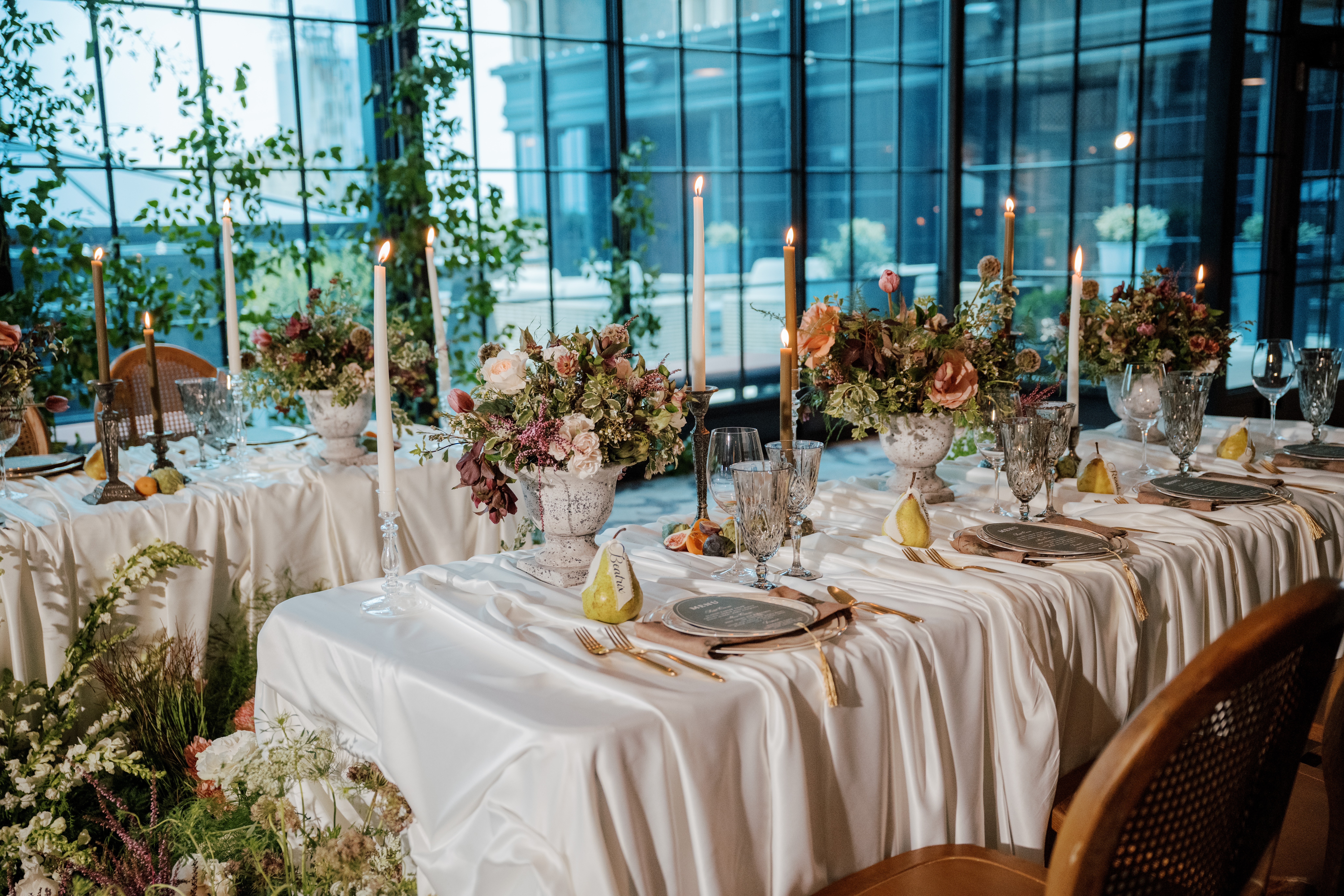 Elegant banquet table with floral centerpieces, candles, and place settings in a glass-walled venue.