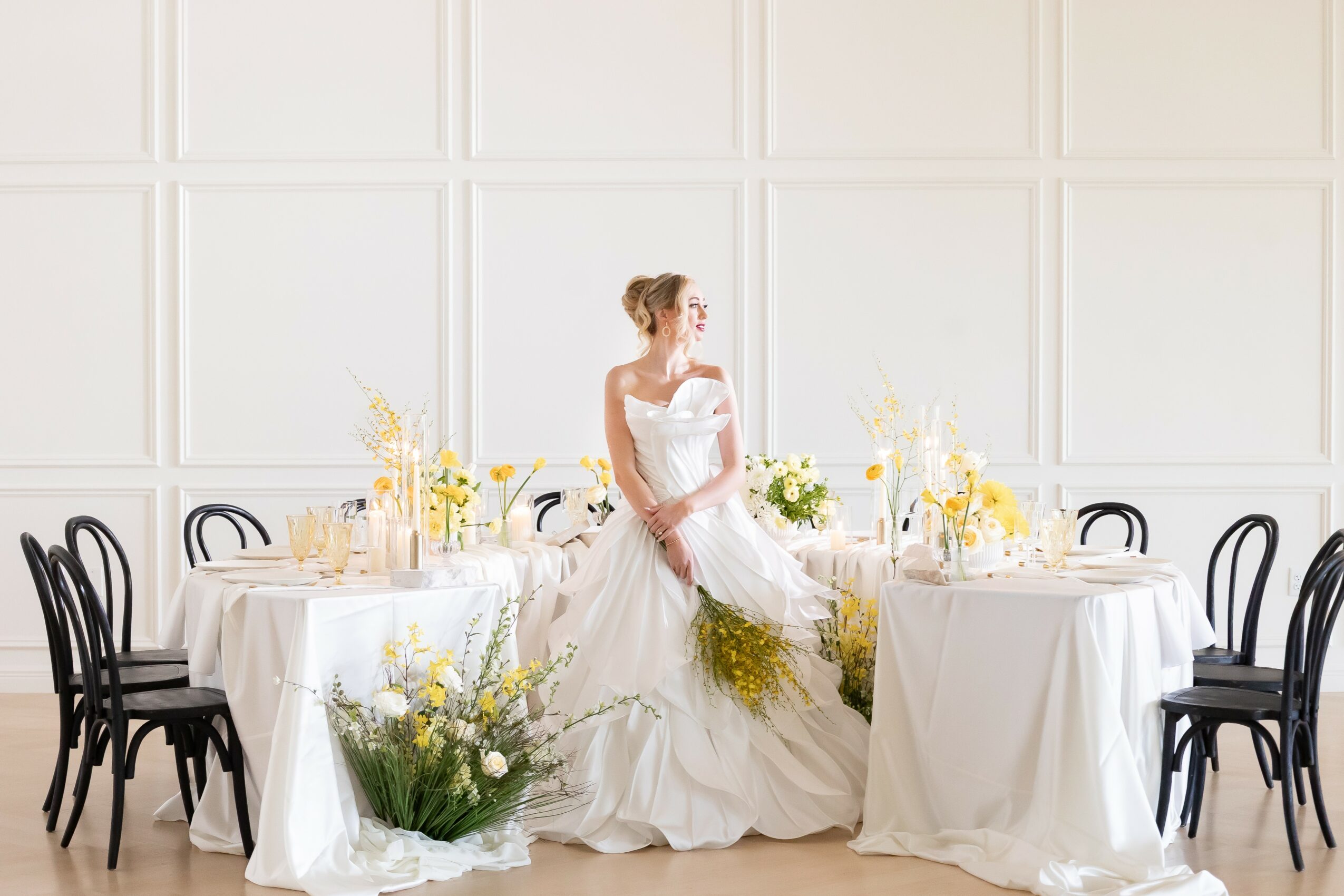 A woman in a wedding dress sits on a table in a decorated dining room with white walls and black chairs.