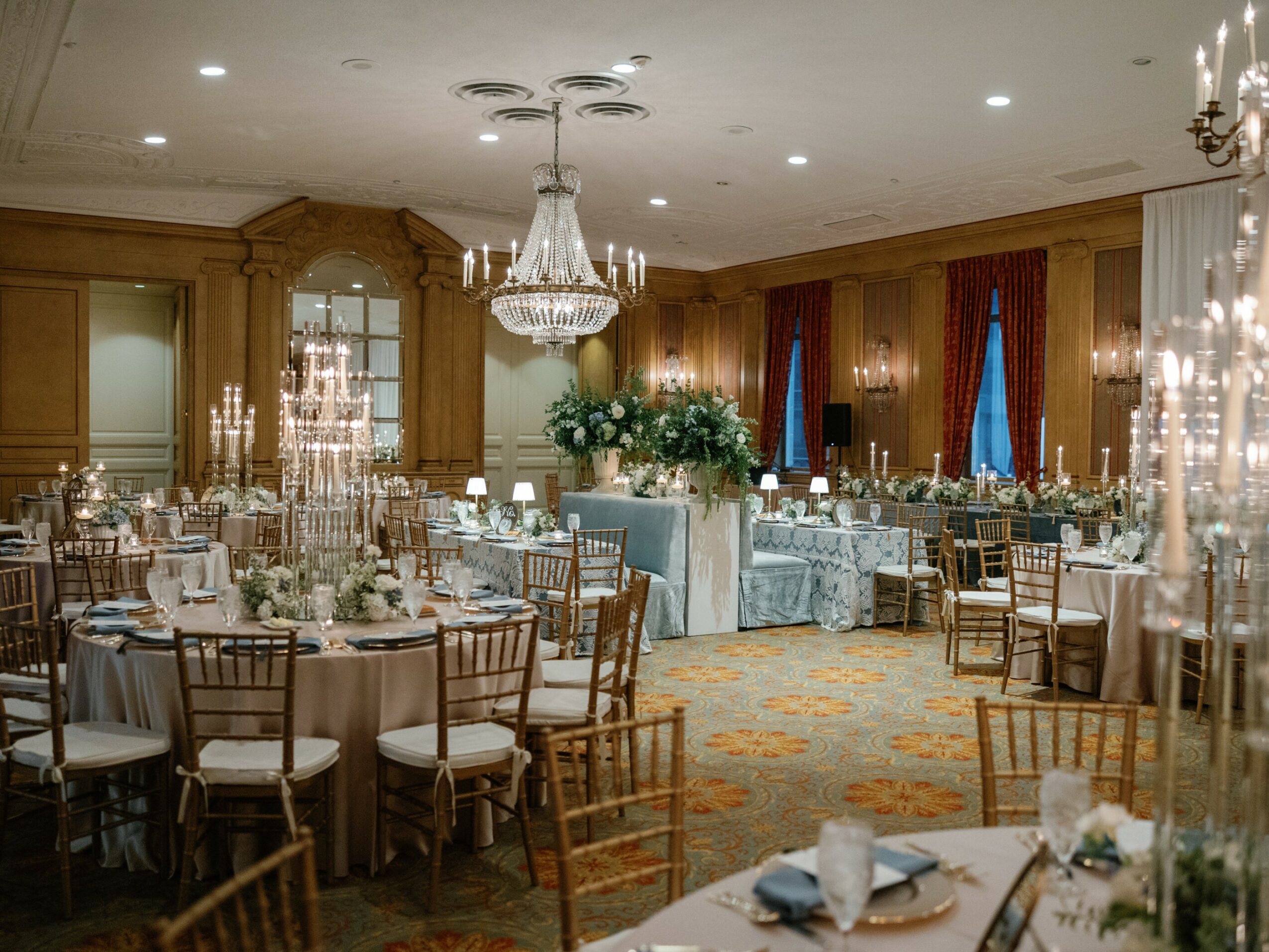 Elegant banquet hall with chandeliers, round tables, and floral centerpieces, decorated for a formal event with wood-paneled walls and red curtains.