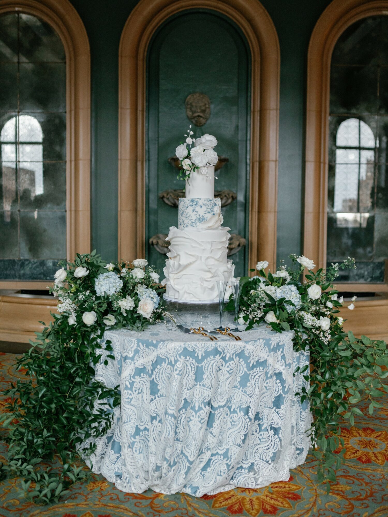 Wedding cake with flowers on a decorated table in front of a mirror, with windows reflected in the background.