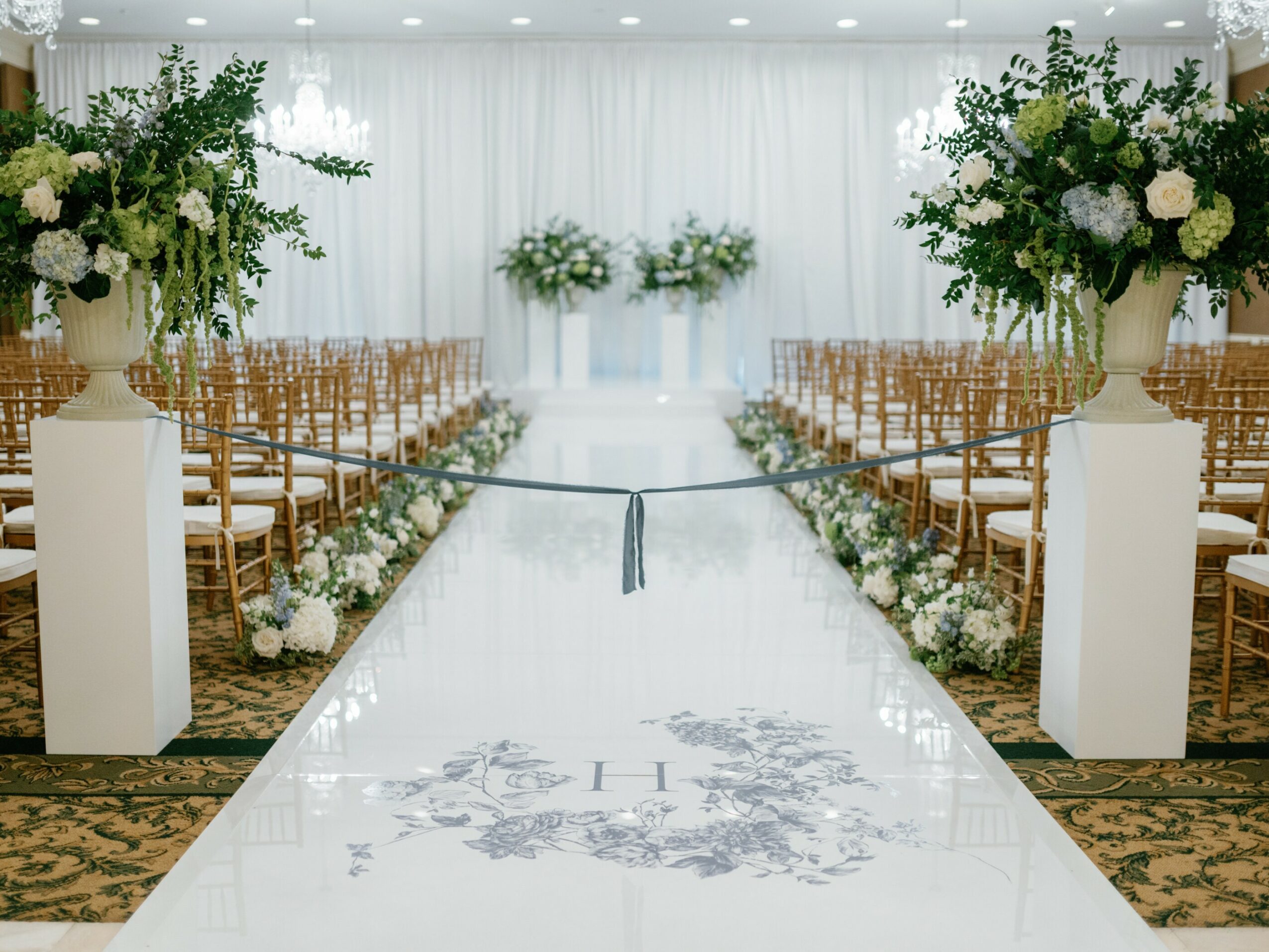 Wedding ceremony setup with rows of chairs, floral arrangements, and a white aisle with a floral design, in a bright room.