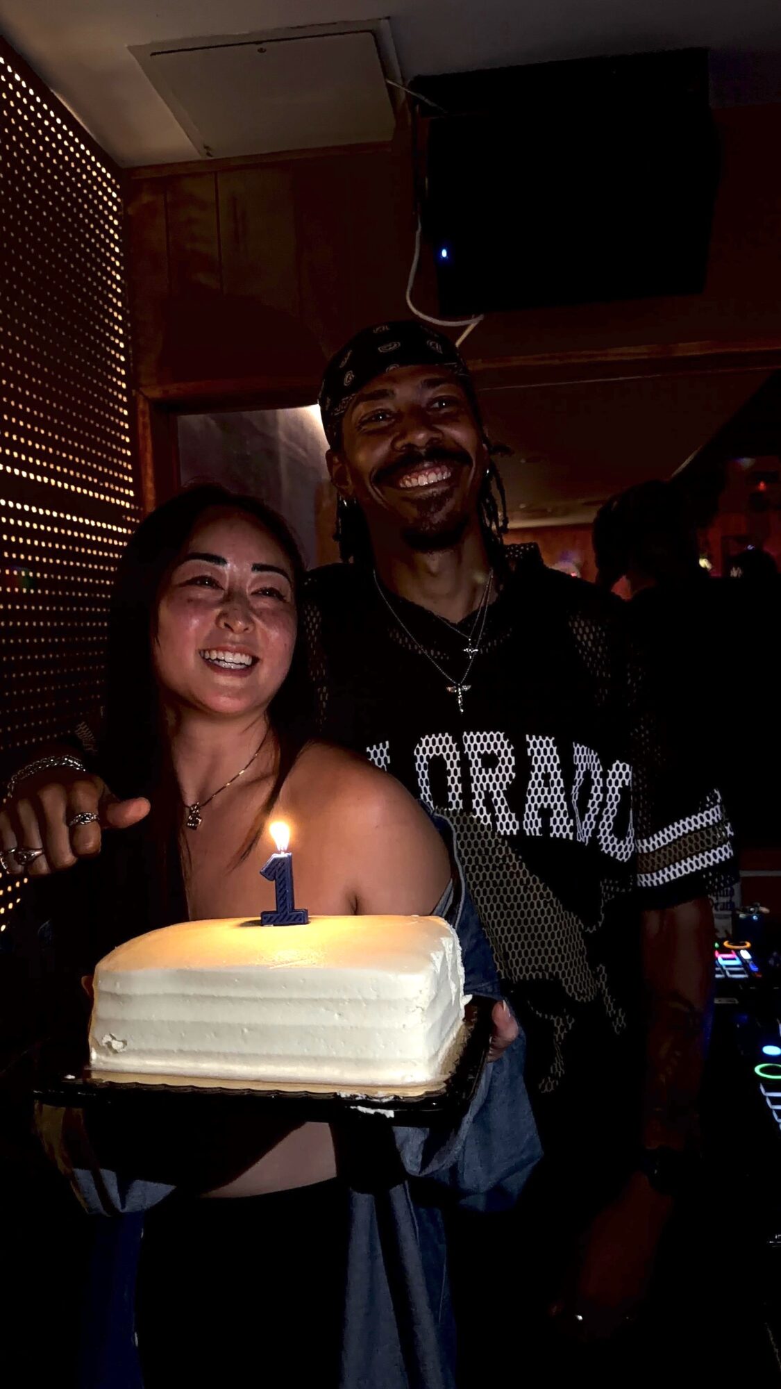 Two people smiling, one holding a birthday cake with a lit candle, in a dimly lit room.