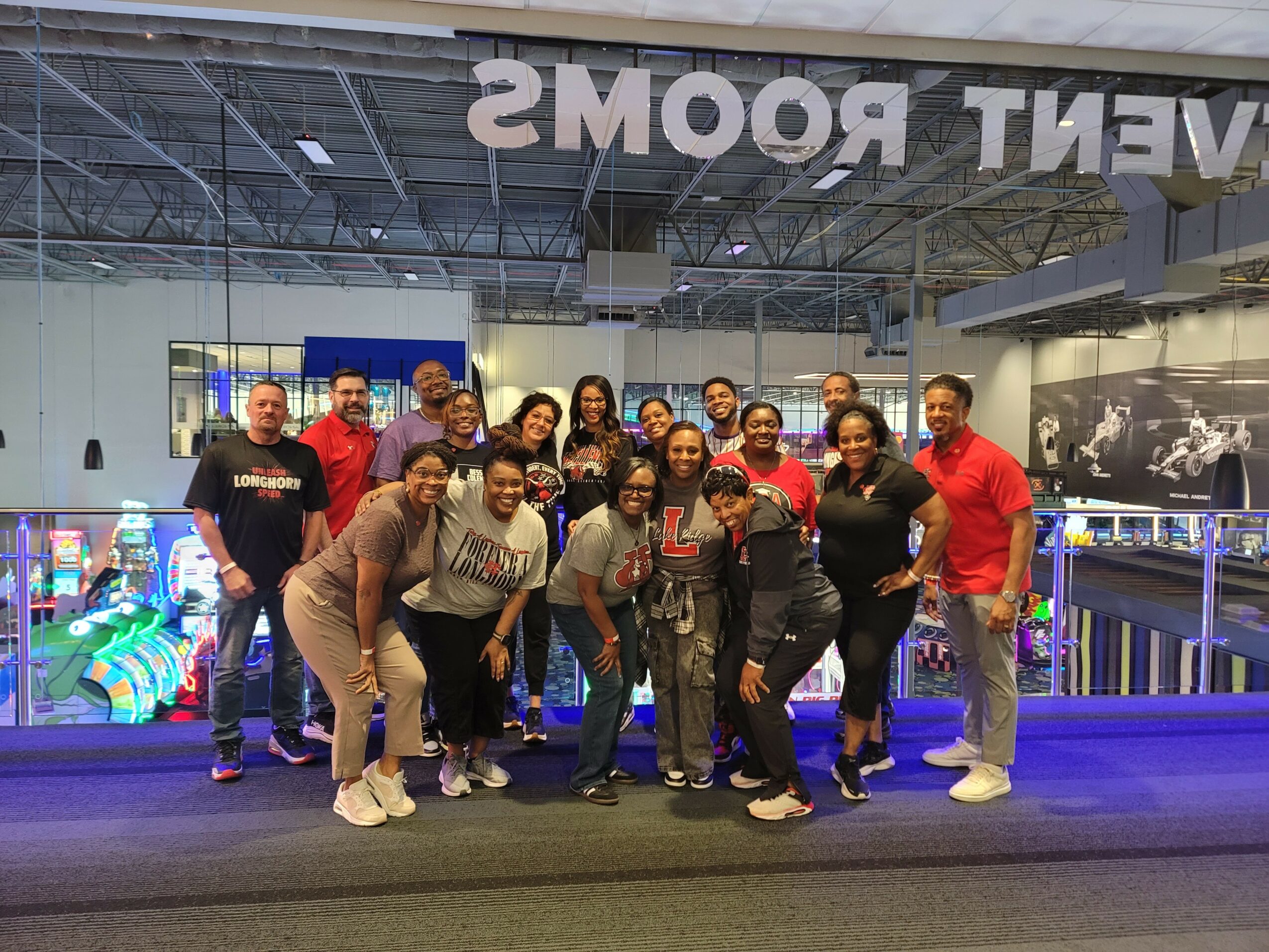 Group of people posing together indoors, some smiling, in front of a glass wall with reflections and displays behind them.