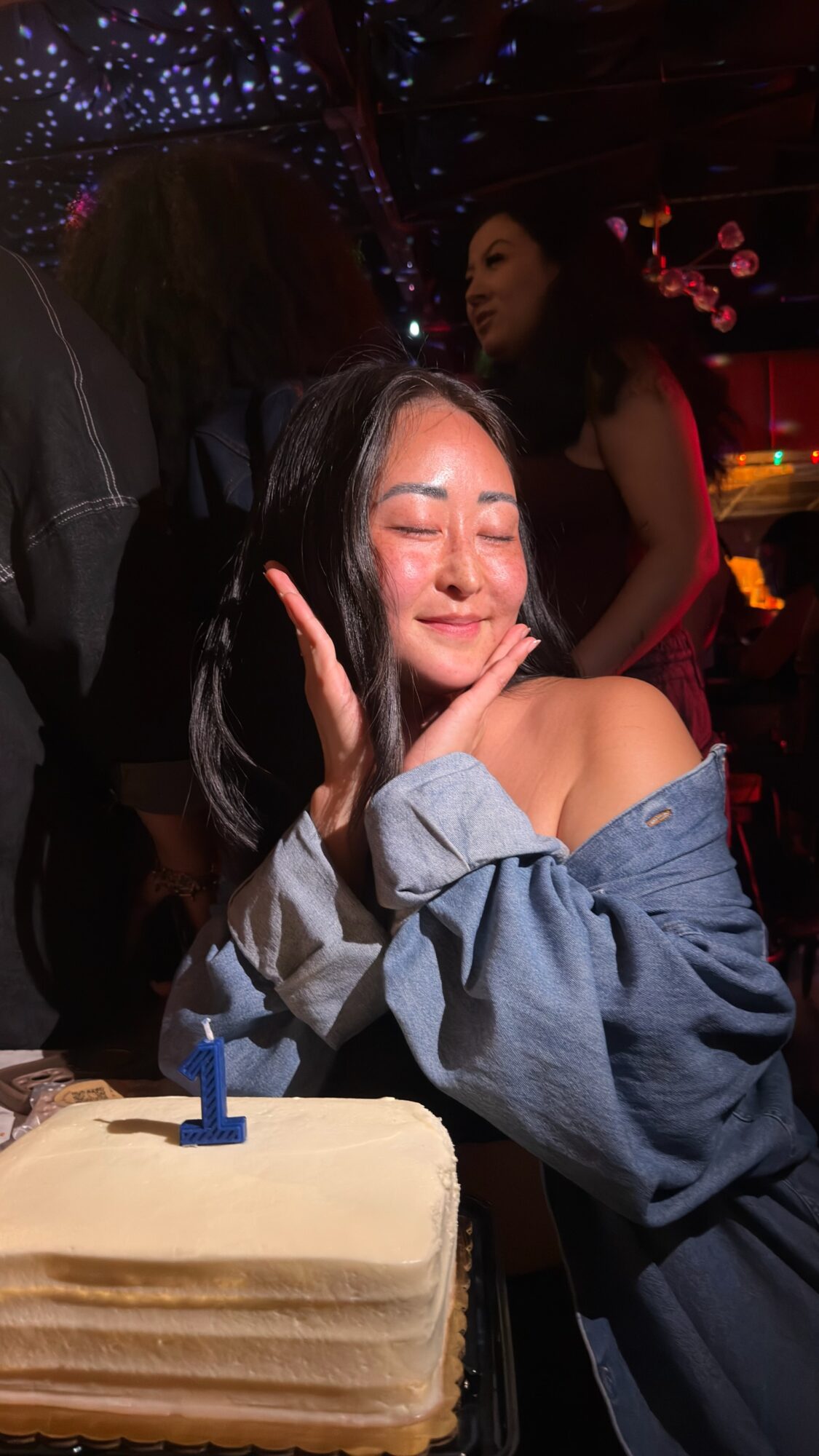 Young woman smiling with eyes closed, resting her face on her hand, in a dimly lit party setting with a cake in front.