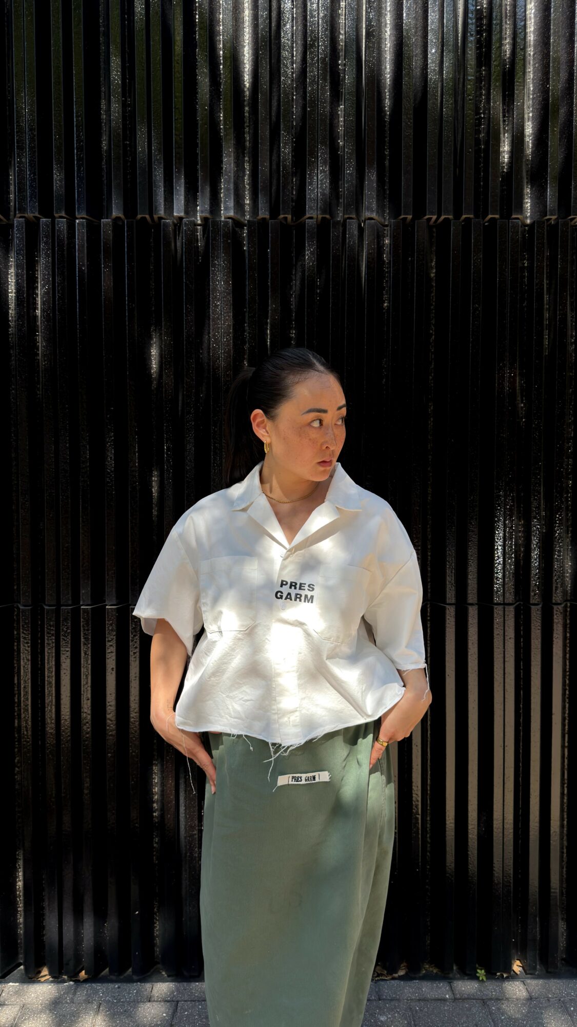 Woman standing in front of a black textured background, wearing a white shirt and green skirt, looking to the side.
