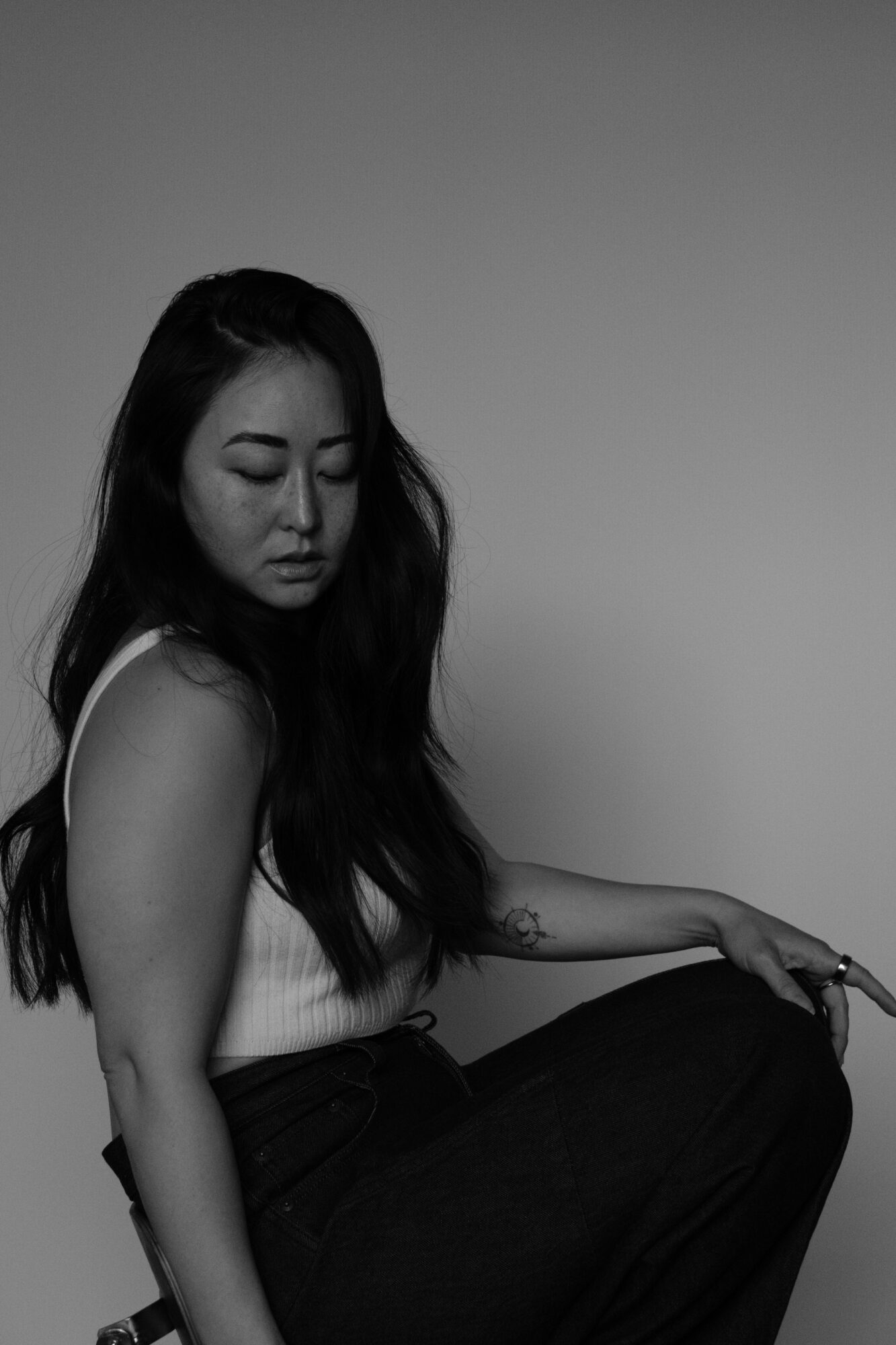 Woman with long hair sitting, looking down, in grayscale, plain background.