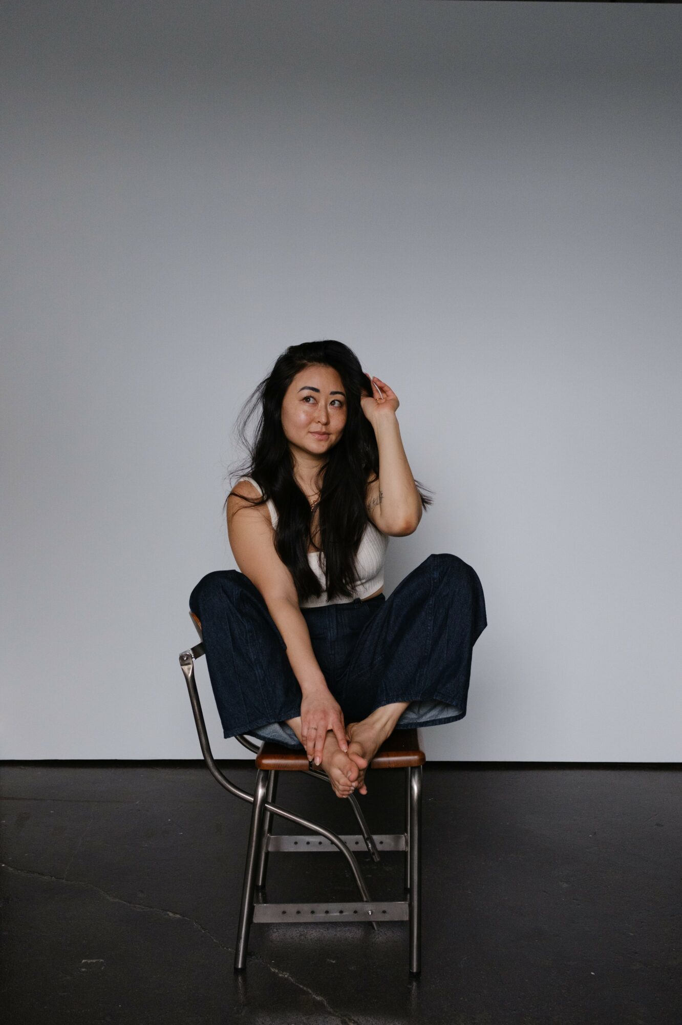 Young woman with long dark hair sitting cross-legged on a chair against a plain light gray background.