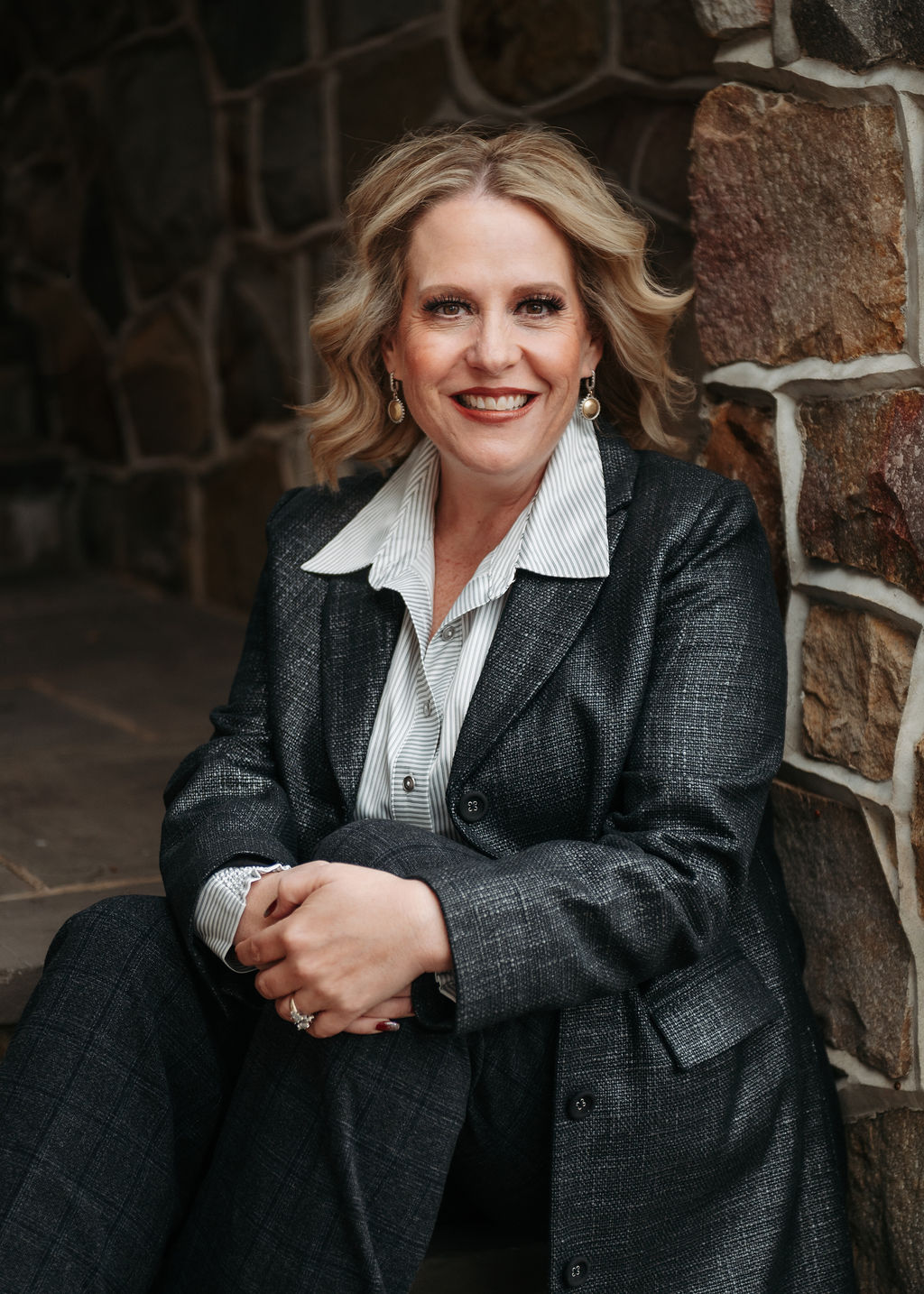 Woman with blonde hair smiling, wearing a dark blazer and white shirt, sitting against a stone wall.