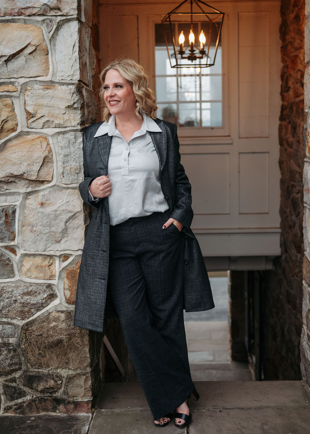 Woman with blonde hair in a coat and pants standing outdoors near a stone wall and door, smiling.