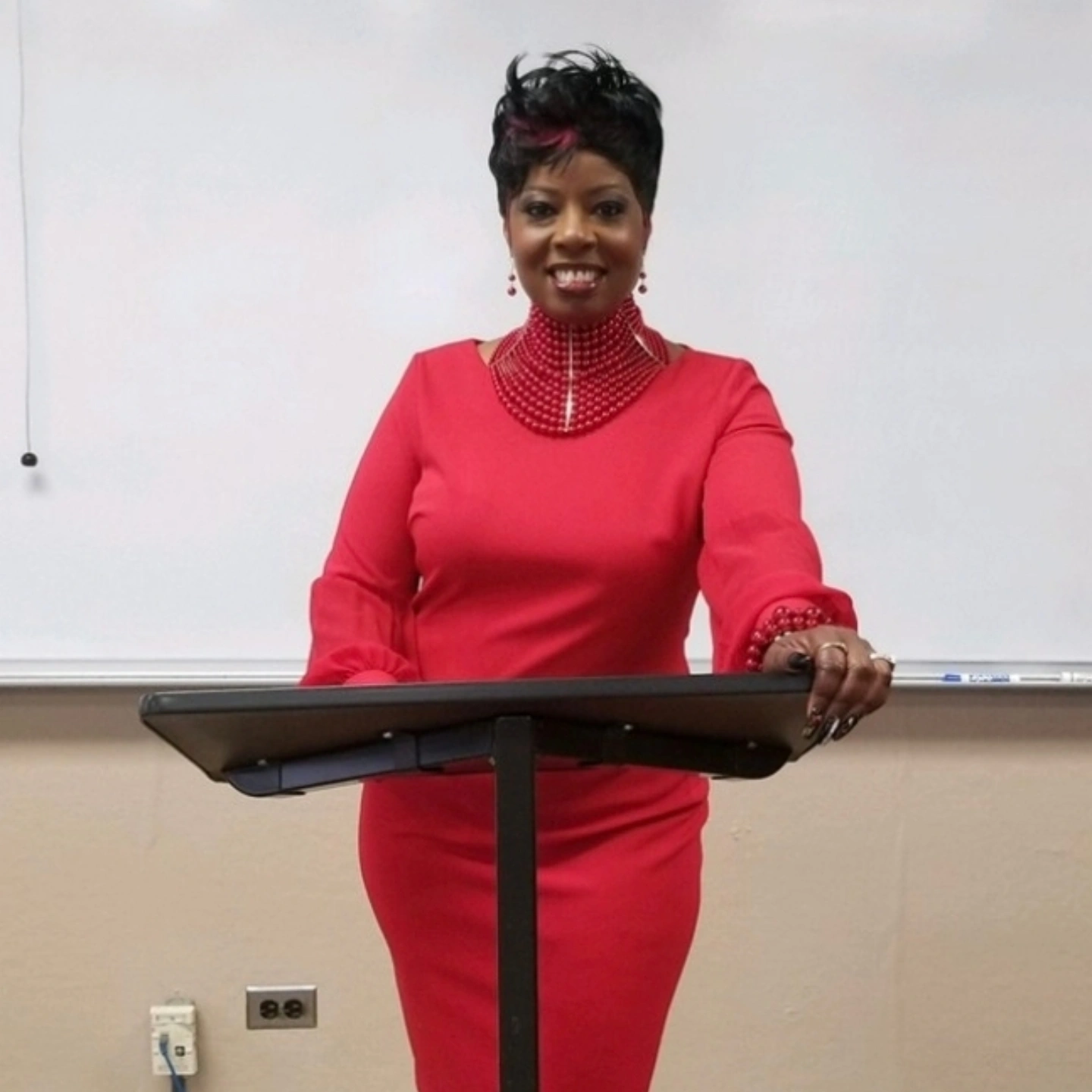 Woman standing at a podium in a red dress, smiling, in front of a whiteboard.
