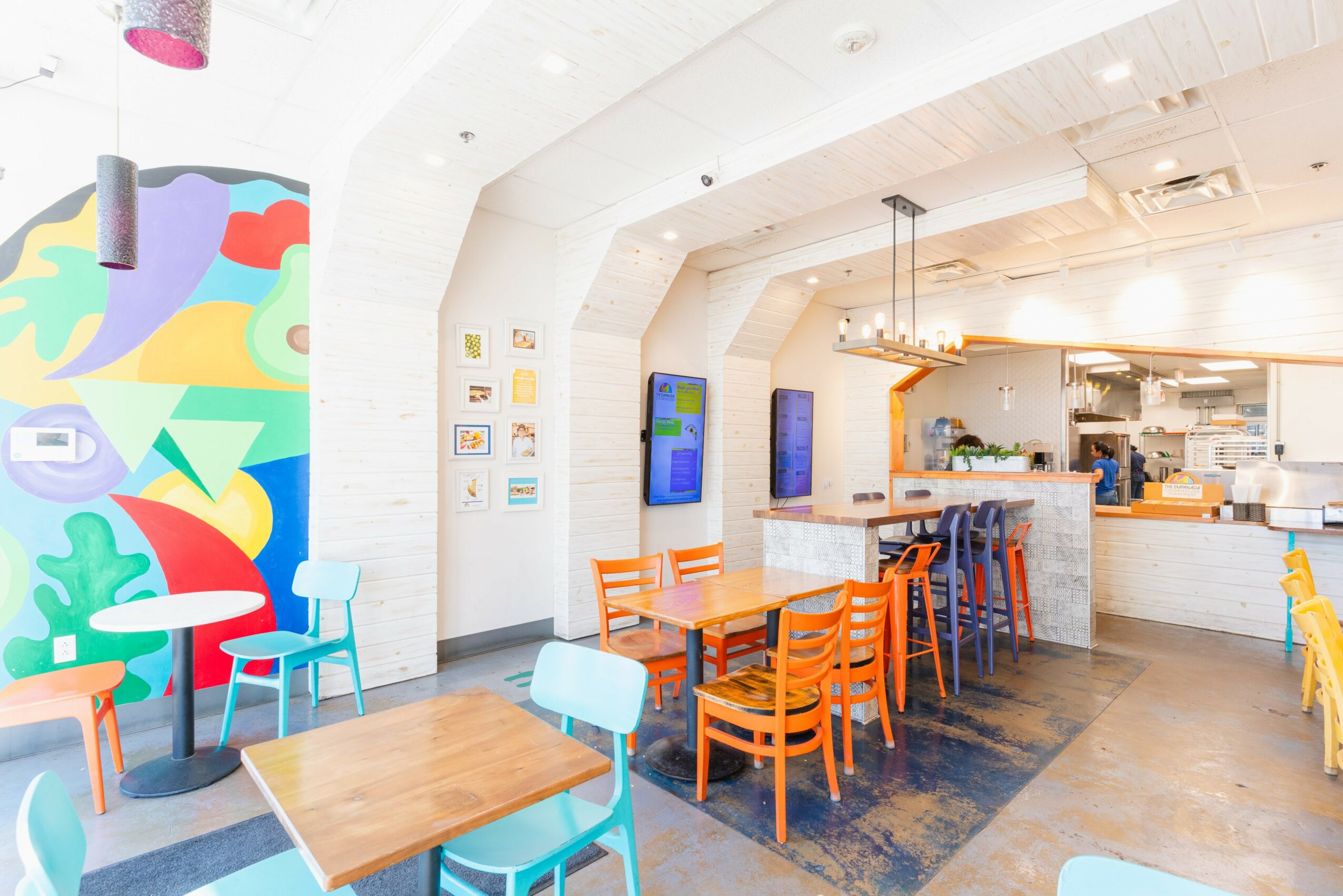 Bright cafe interior with colorful wall art, wooden tables and chairs, and a bar area with stools, modern decor.