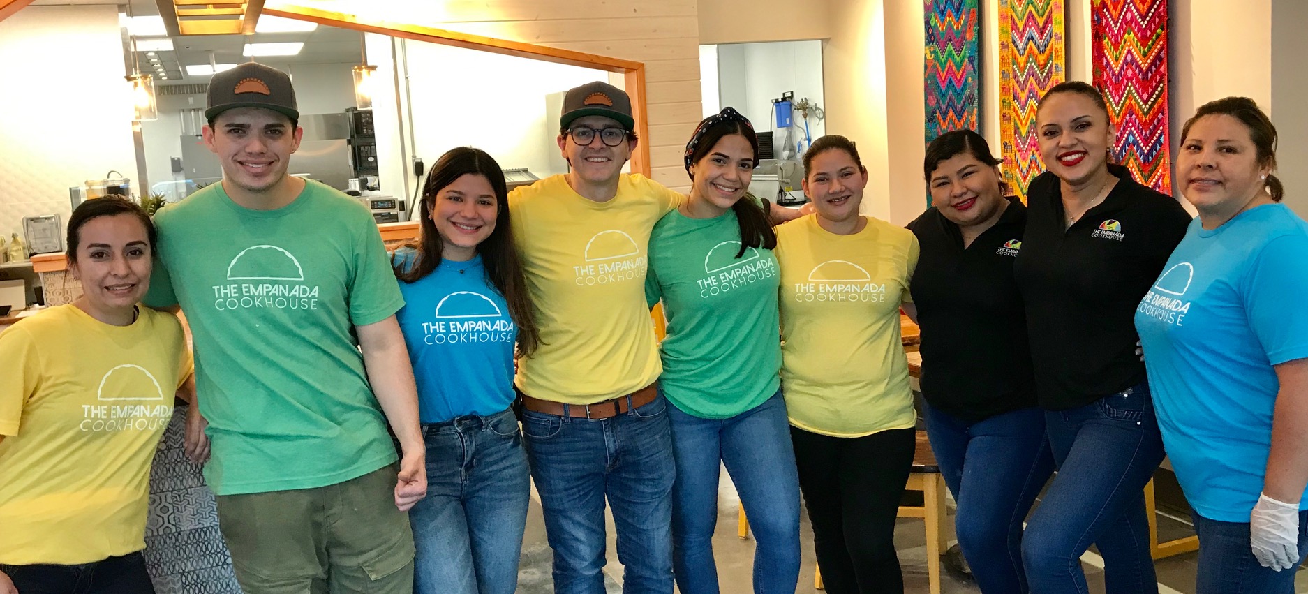 Group of nine people standing indoors, smiling, wearing colorful shirts, with vibrant decorations in the background.