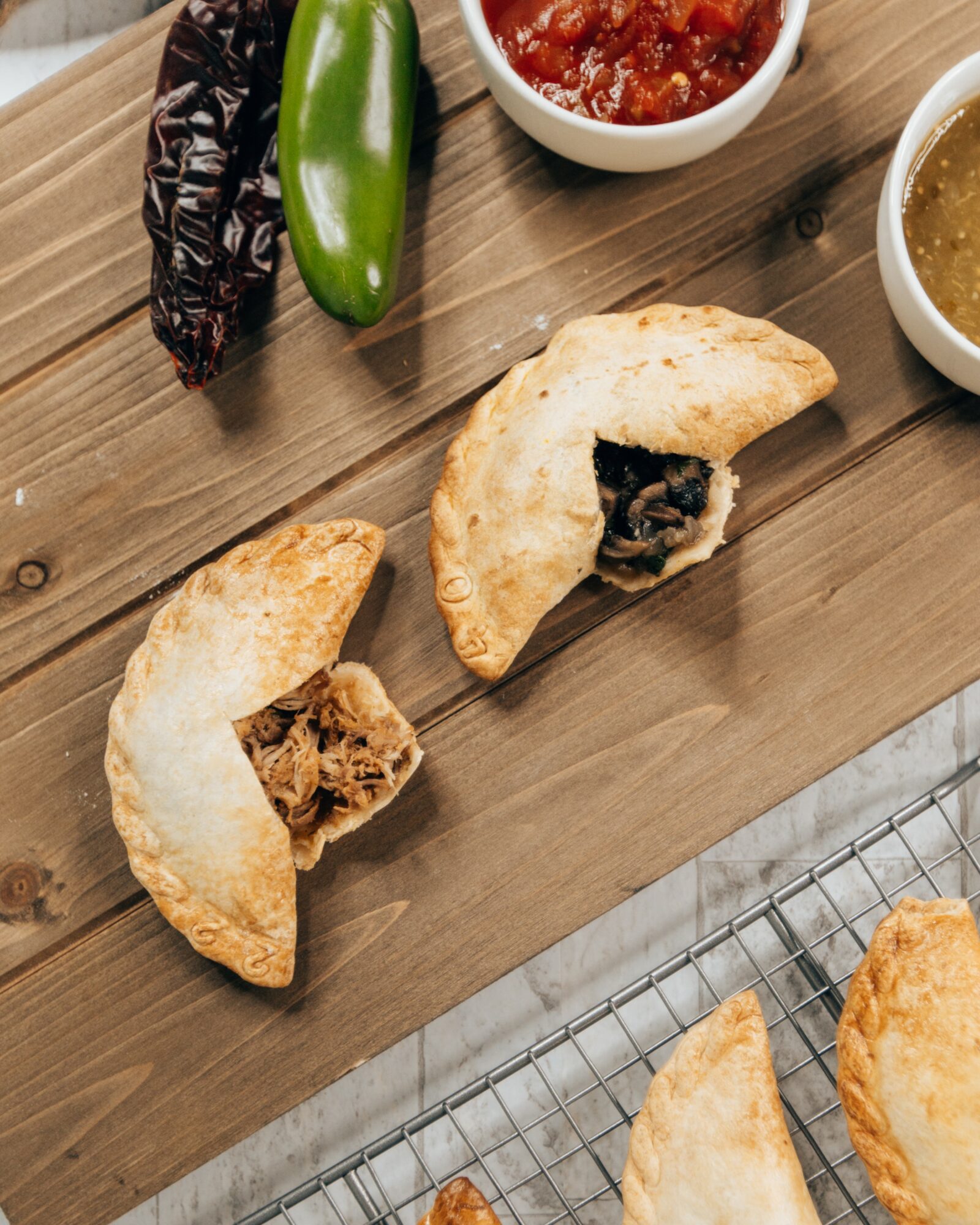 Two empanadas on a wooden surface, one cut open showing filling, with bowls of salsa and sauce nearby.