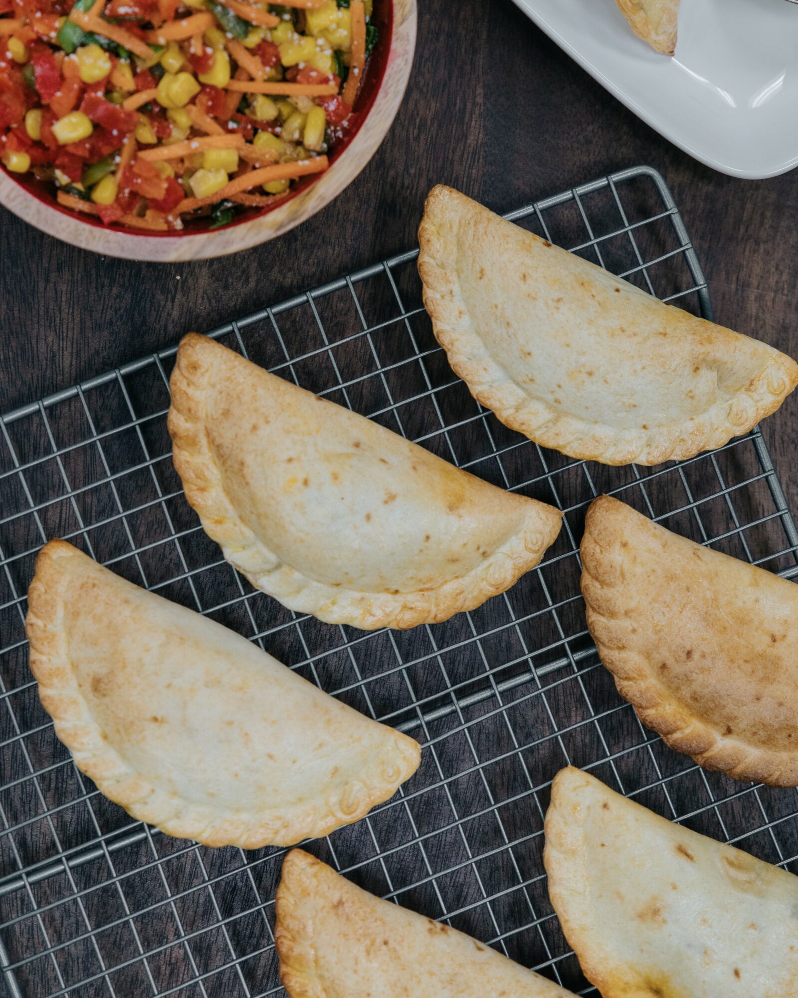 Six fried empanadas on a wire rack with a bowl of salad in the background.