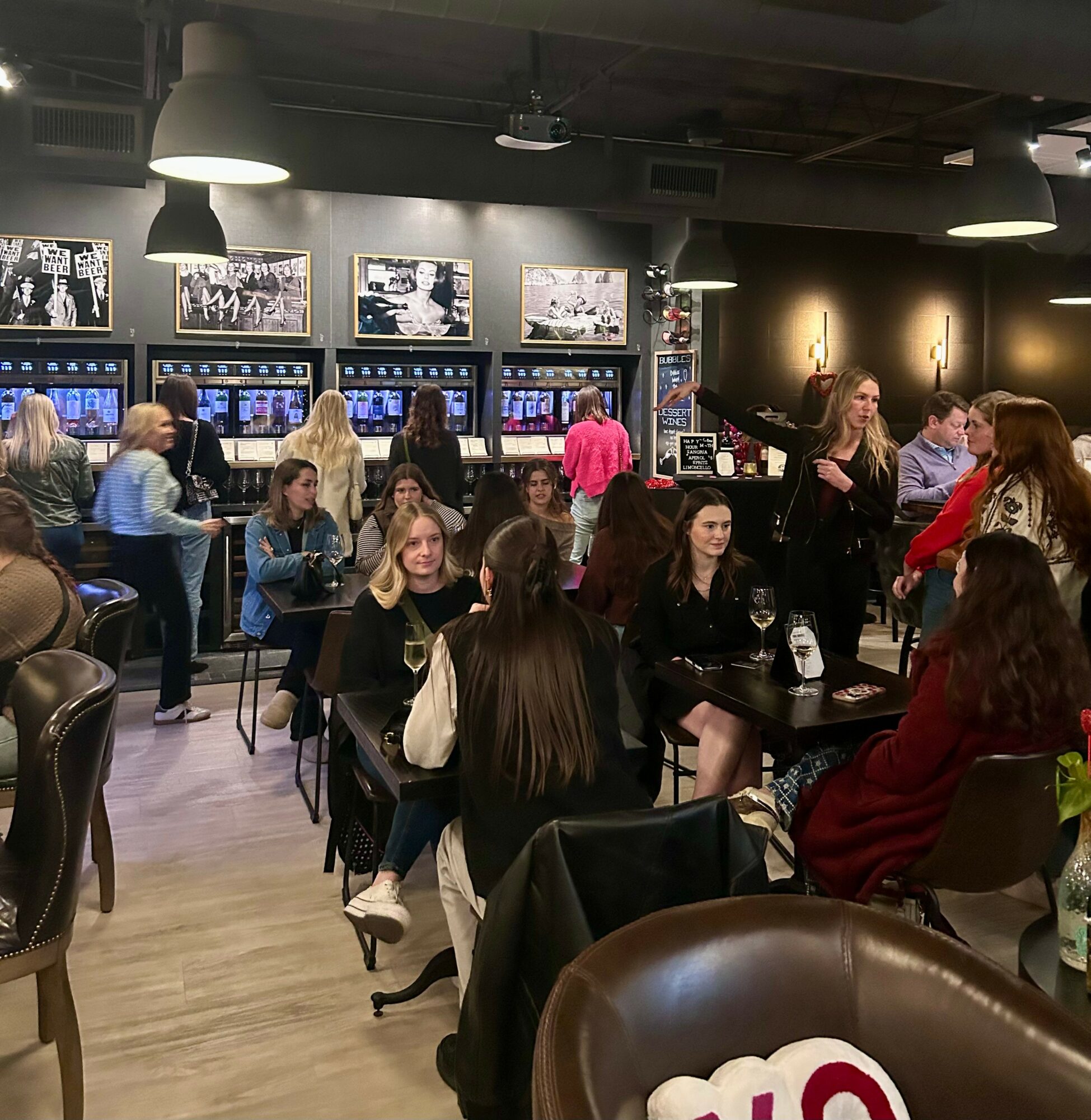 People sitting and standing in a lively bar or restaurant, with gaming machines and framed pictures on the wall.