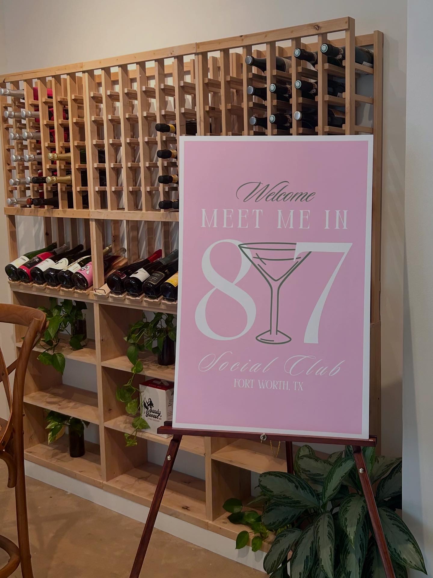 Wine rack with bottles and a pink sign on a stand, decorated with plants, in a room.