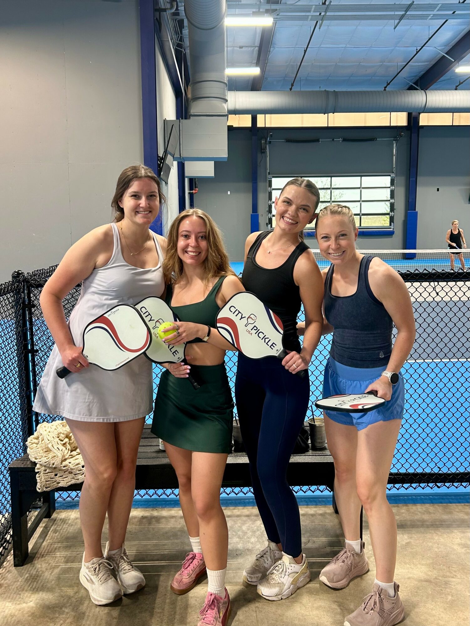 Four women standing together inside a sports facility, holding tennis rackets and a paddle, smiling at the camera.