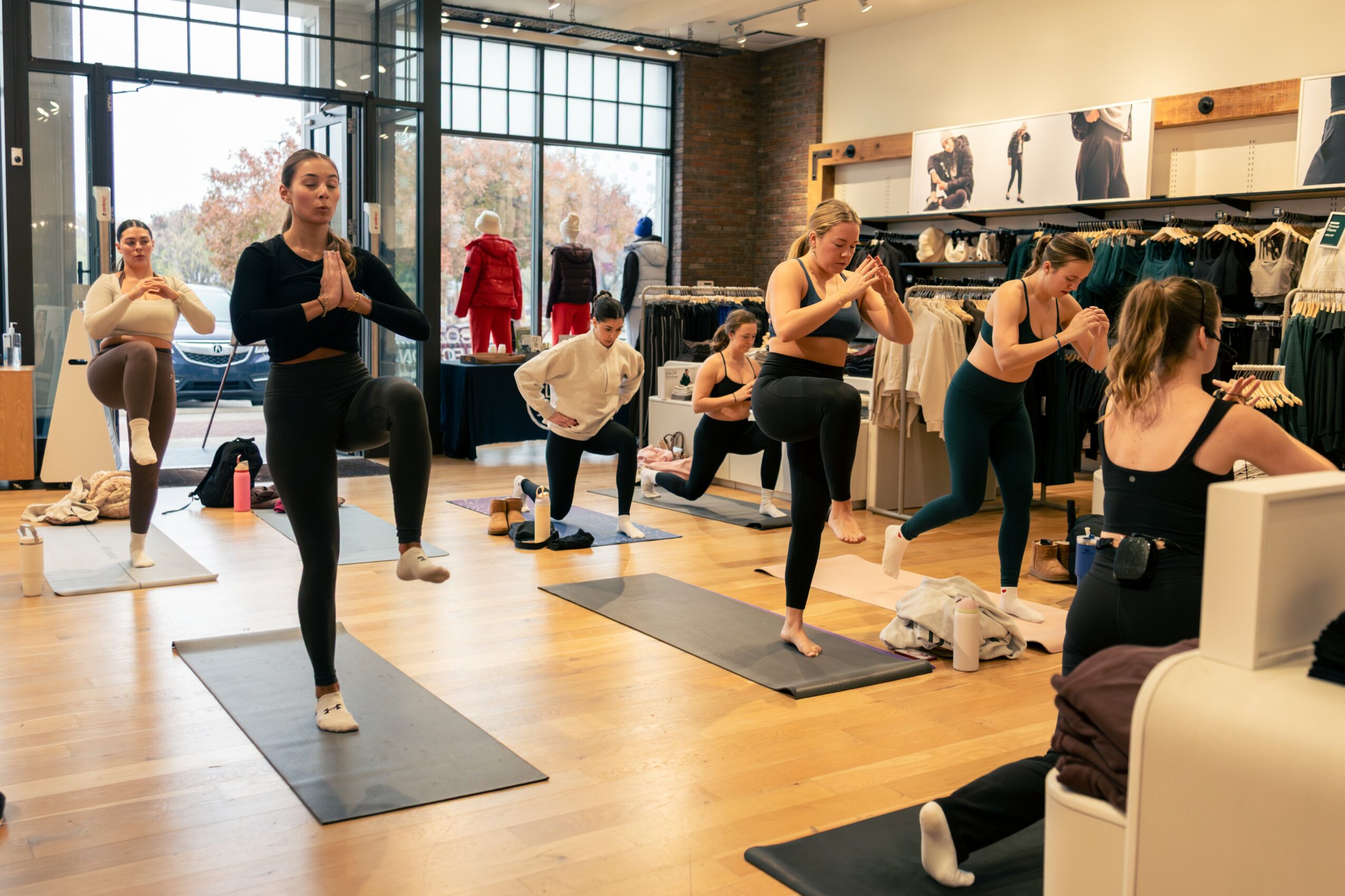 Group of women practicing yoga in a studio with large windows and clothing displays, some in tree pose, others stretching.