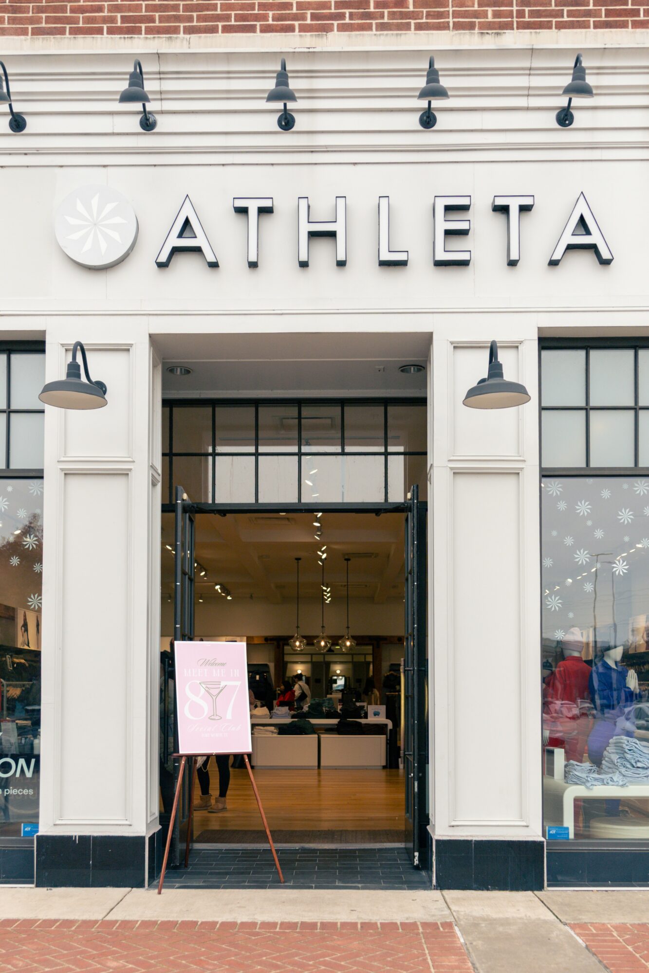 Storefront with sign reading 'ATHLETA' and open entrance, two black lamps on white columns, and a pink sign outside.