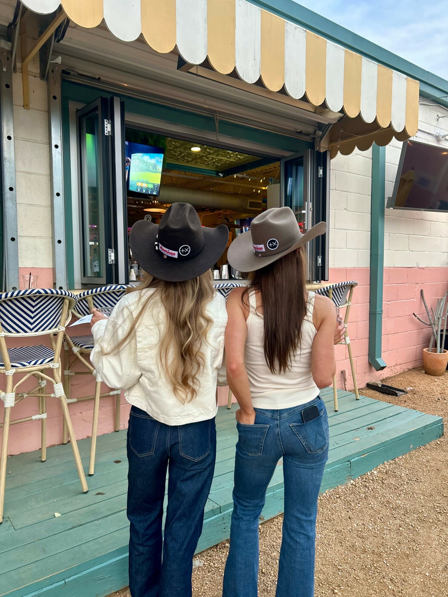 Two women with long hair wearing hats and jeans stand outside a colorful shop with a striped awning.