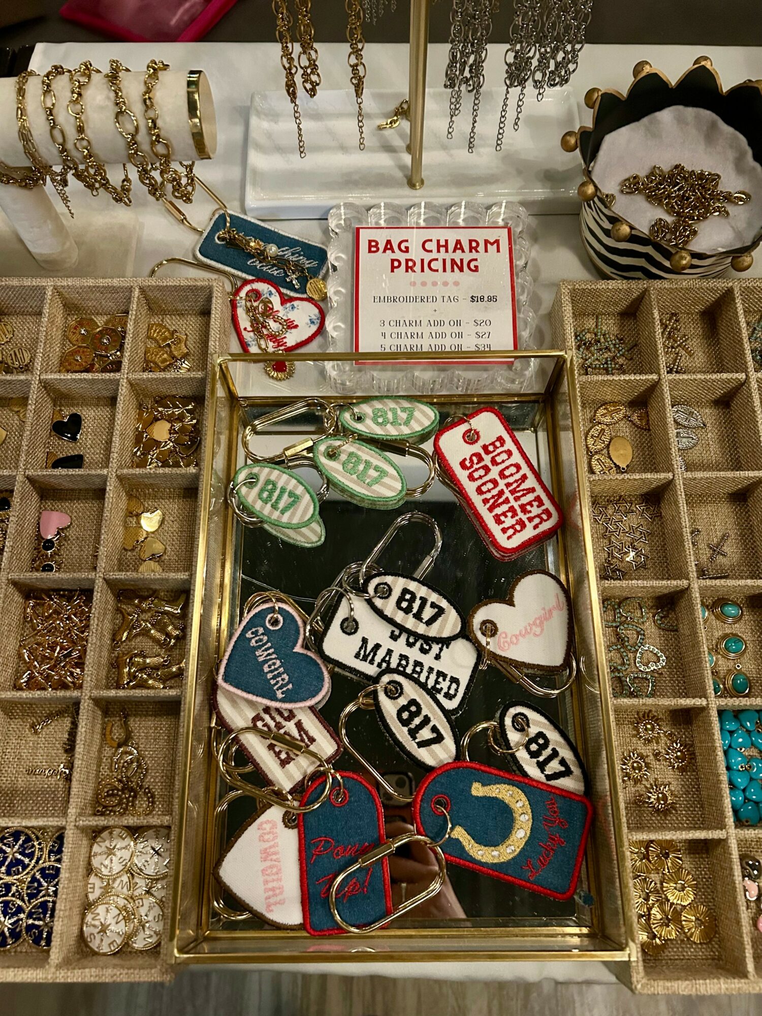 Display of jewelry and bag charms with a sign reading 'Bag Charm Pricing' in a jewelry store, various charms and accessories visible.