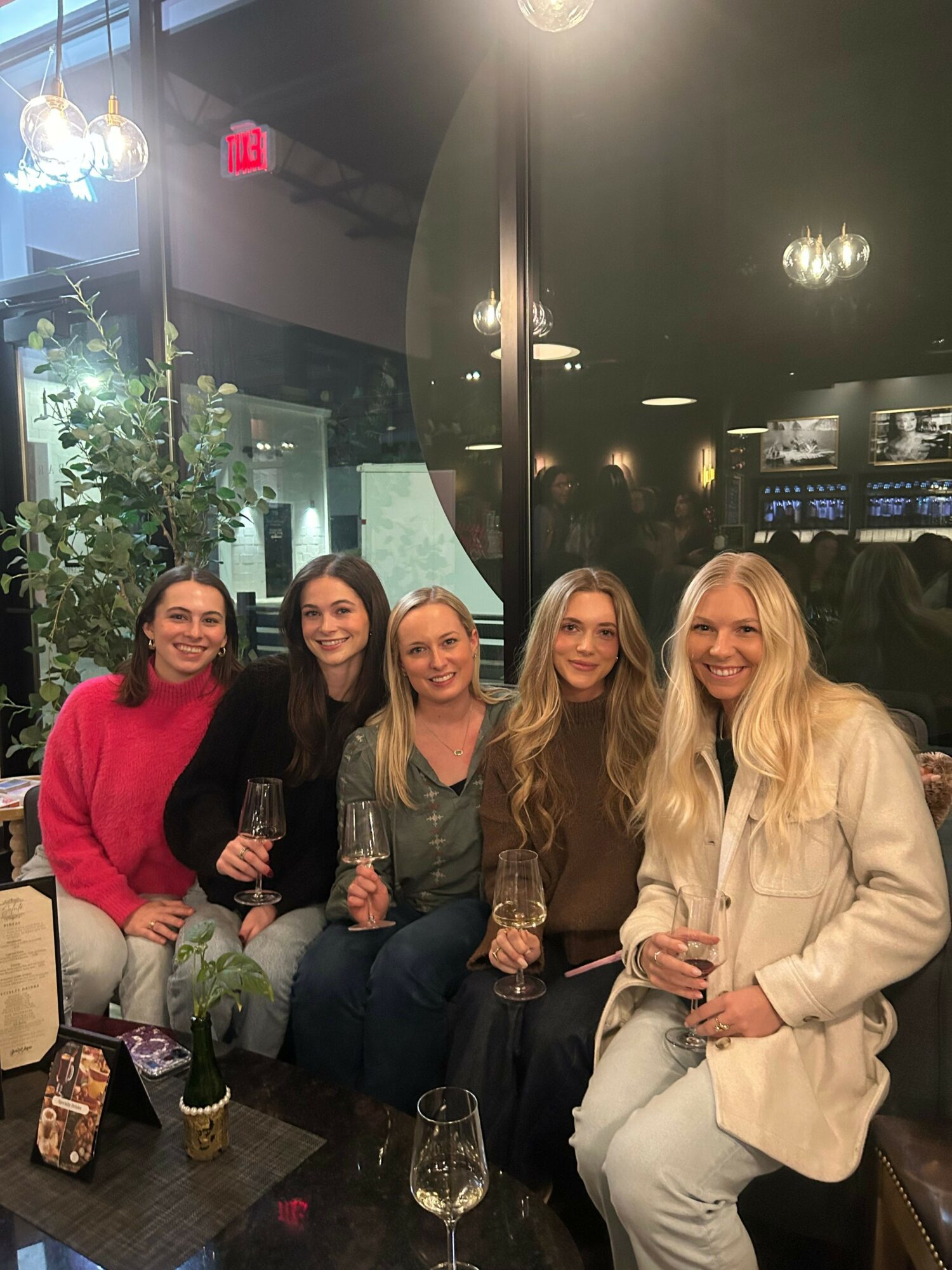 Five women sitting together in a dimly lit indoor space, smiling and holding drinks, with a window and plants in the background.