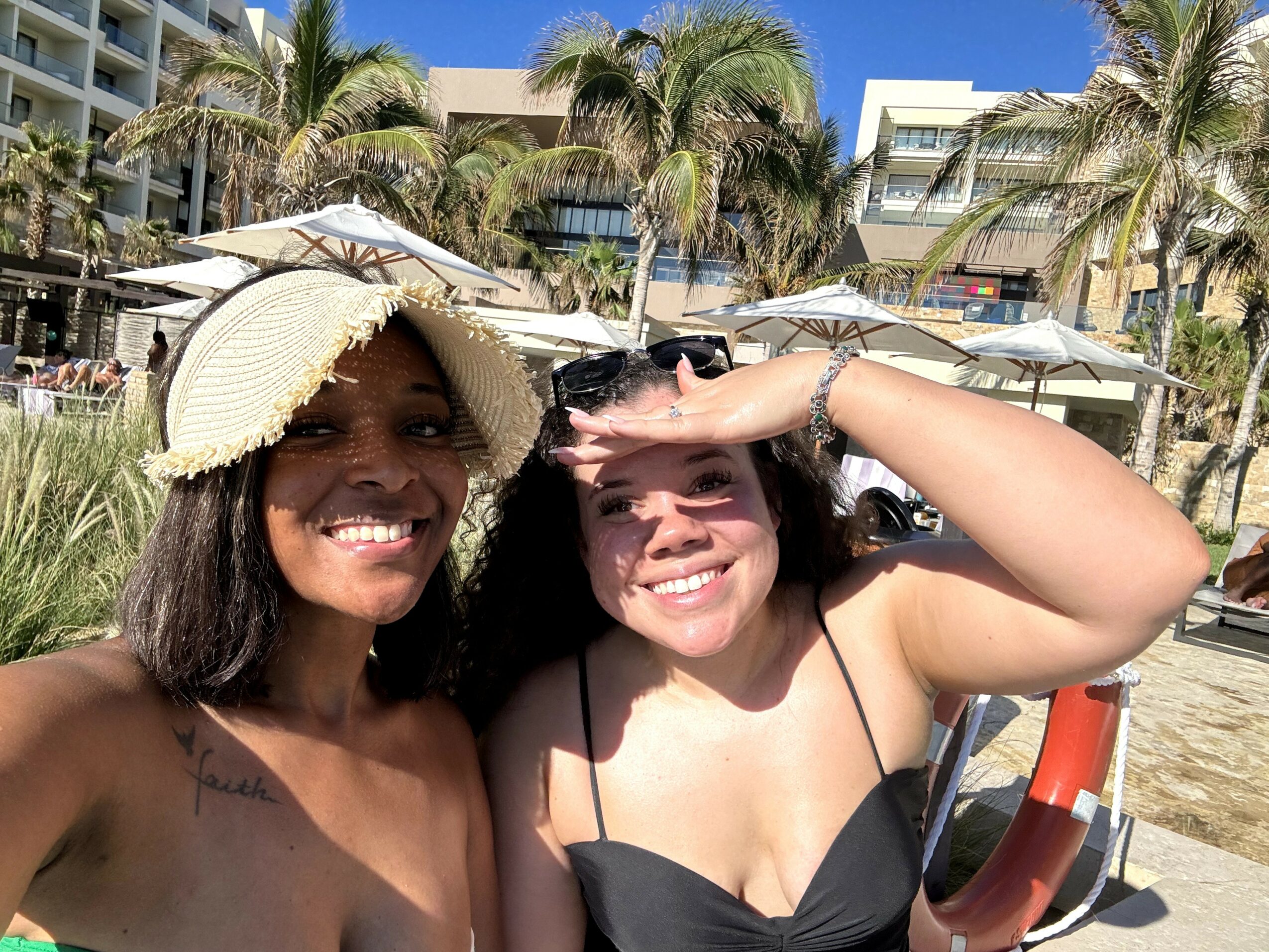 Two women smiling outdoors with palm trees and buildings in background, one wearing a straw hat, the other shielding eyes from sun.
