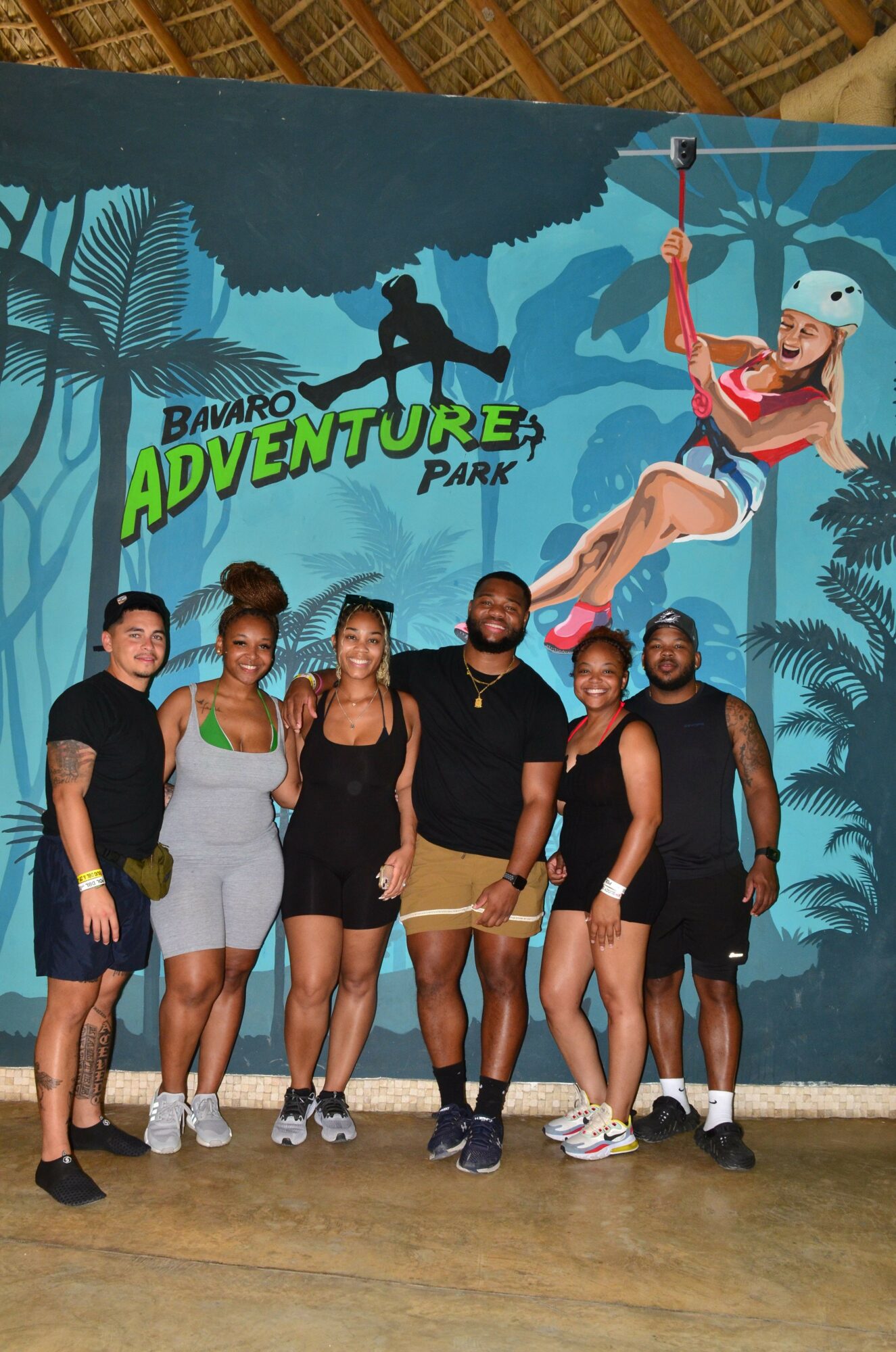 Group of seven people standing in front of a colorful adventure park backdrop, some holding a flag, smiling.