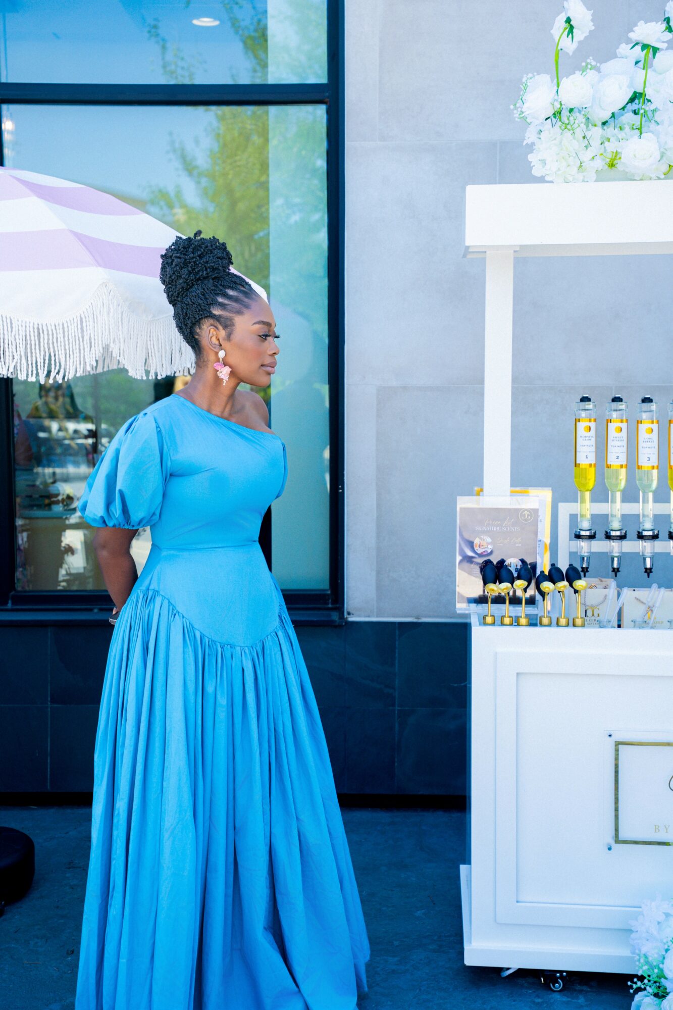 Woman in blue dress standing outside near a white display with bottles, glass window reflecting trees, and a white umbrella.