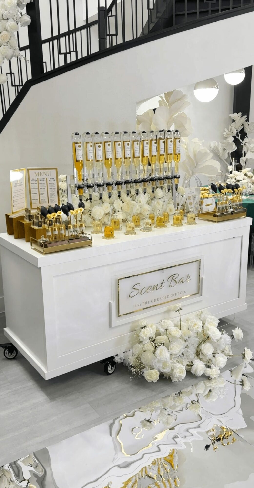 Display table with drinks, flowers, and decorations, set against a white wall with a mirror and staircase, decorated with white flowers.