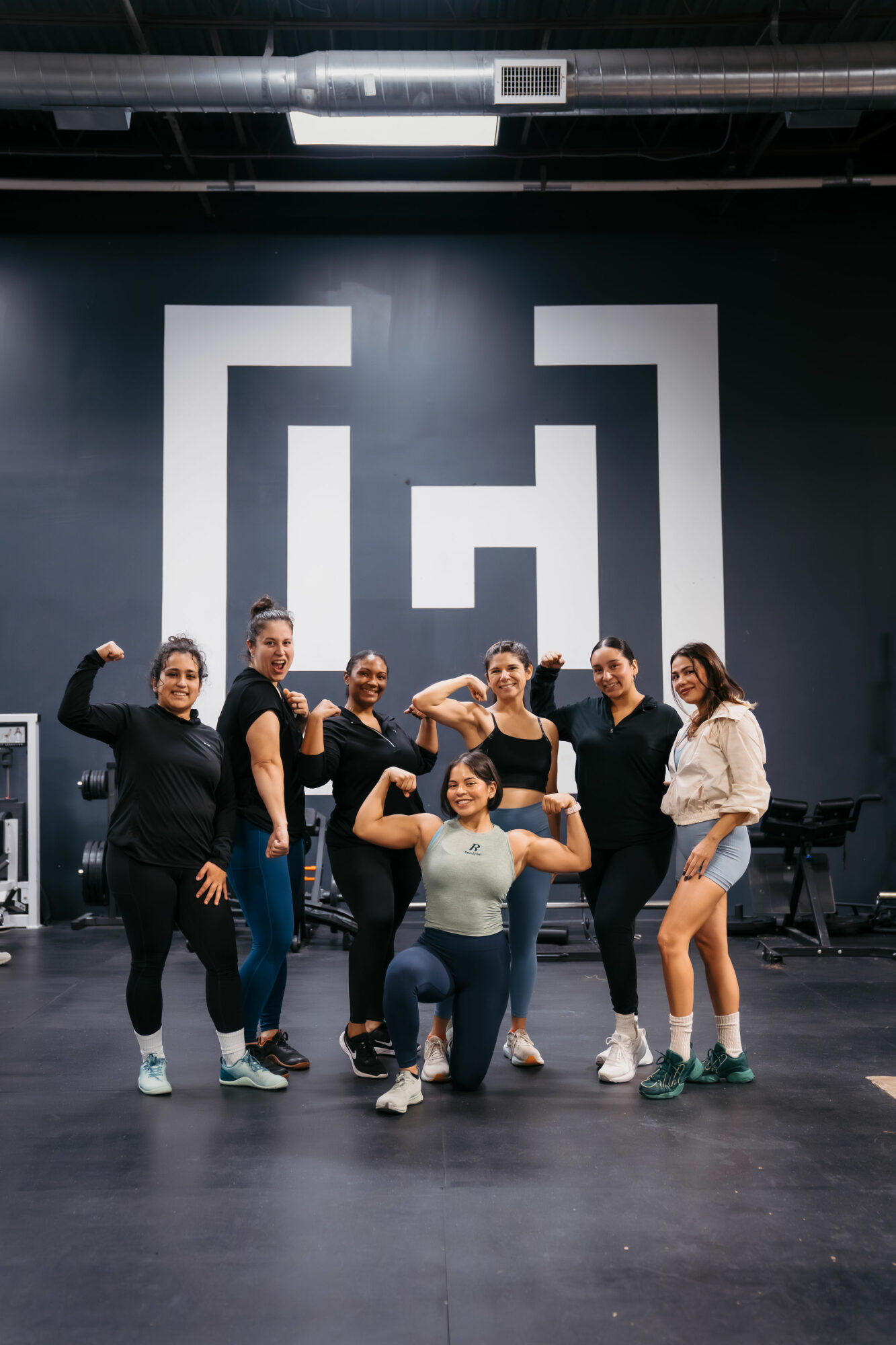Group of seven women posing in a dance studio with a large logo on the wall behind them, some flexing muscles.