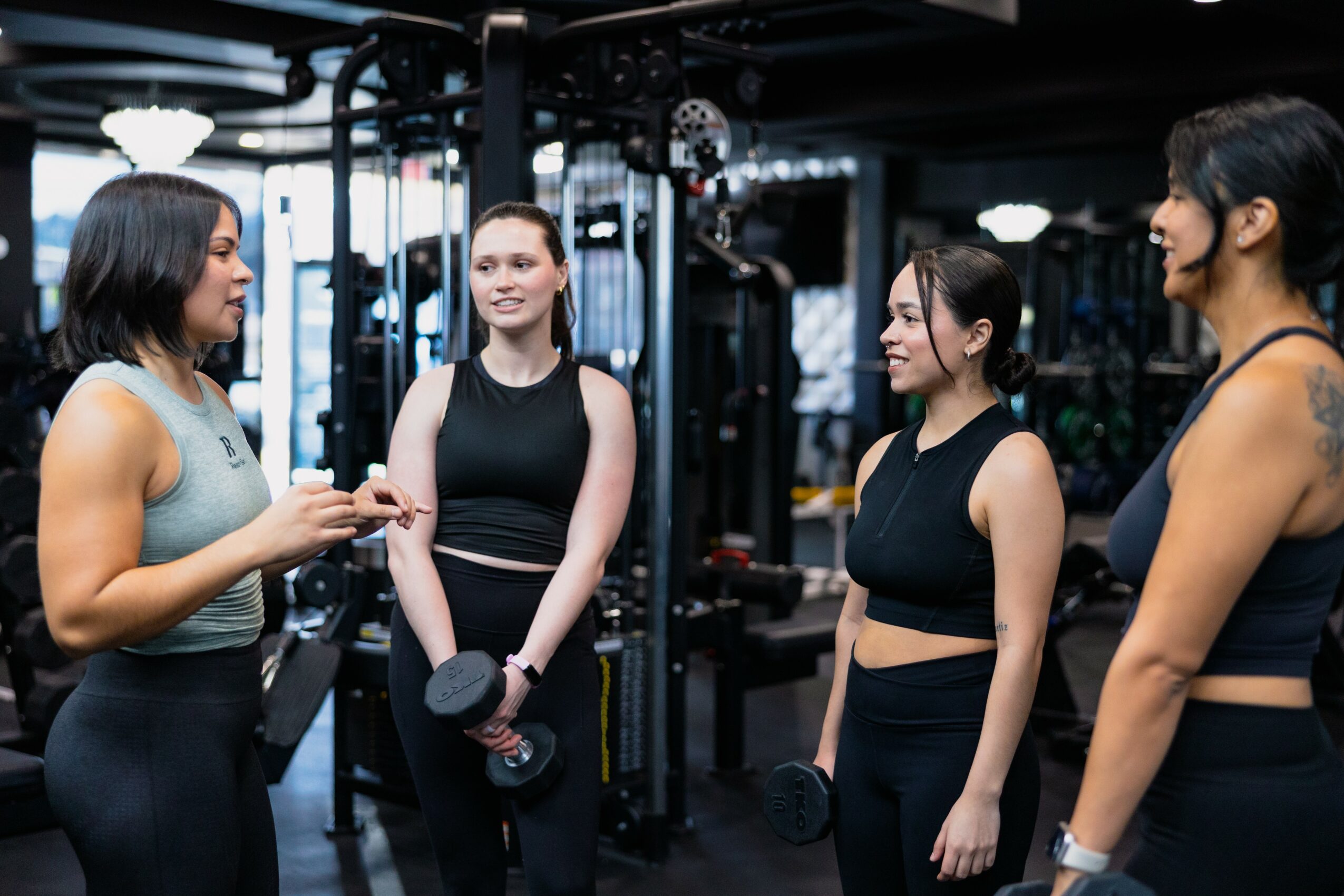 Four women in workout clothes talking in a gym, holding dumbbells, with gym equipment in the background.