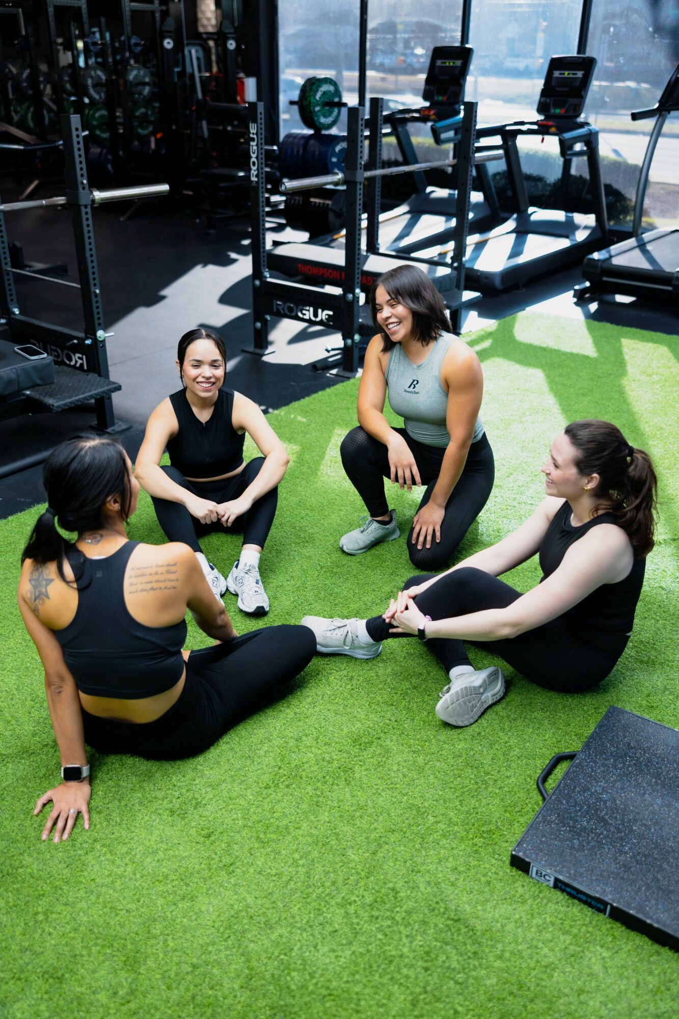 Four women sitting on green artificial turf in a gym, talking and smiling, with exercise equipment in the background.