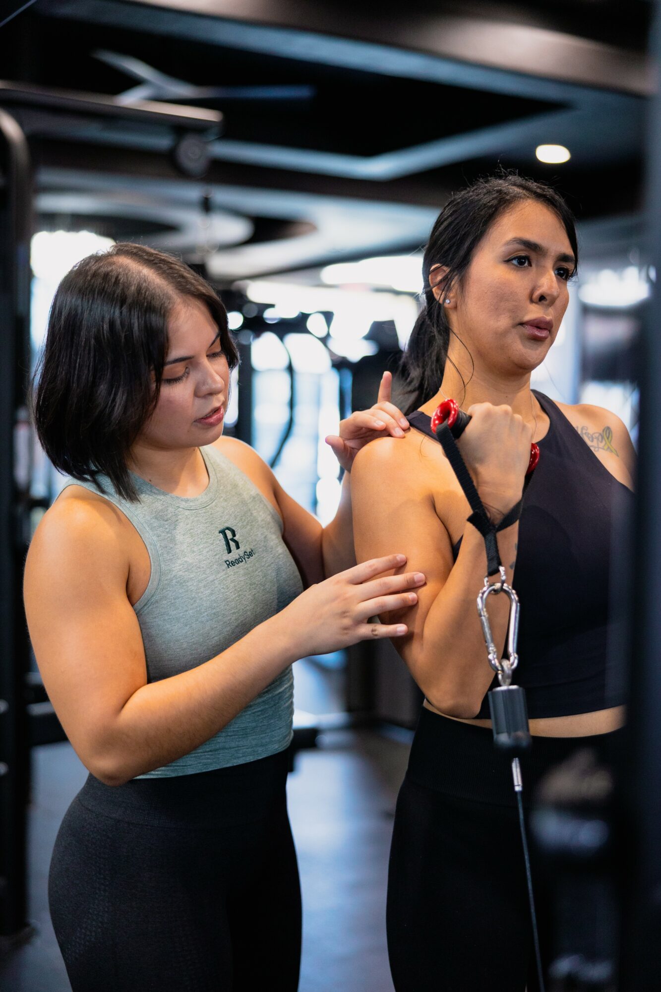Two women in a gym, one adjusting the other's arm, with gym equipment in the background.
