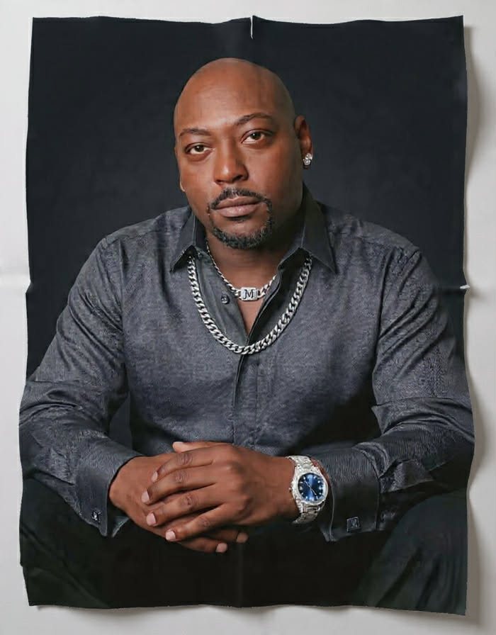 Portrait of a man with a serious expression, wearing a dark shirt, jewelry, and a watch, sitting with hands clasped.
