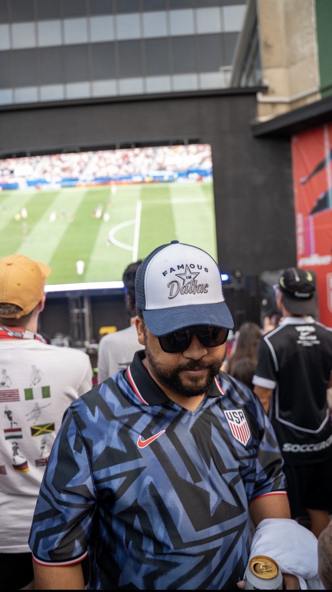 Man wearing sunglasses, a cap, and a sports jersey watching a soccer game on a large screen.