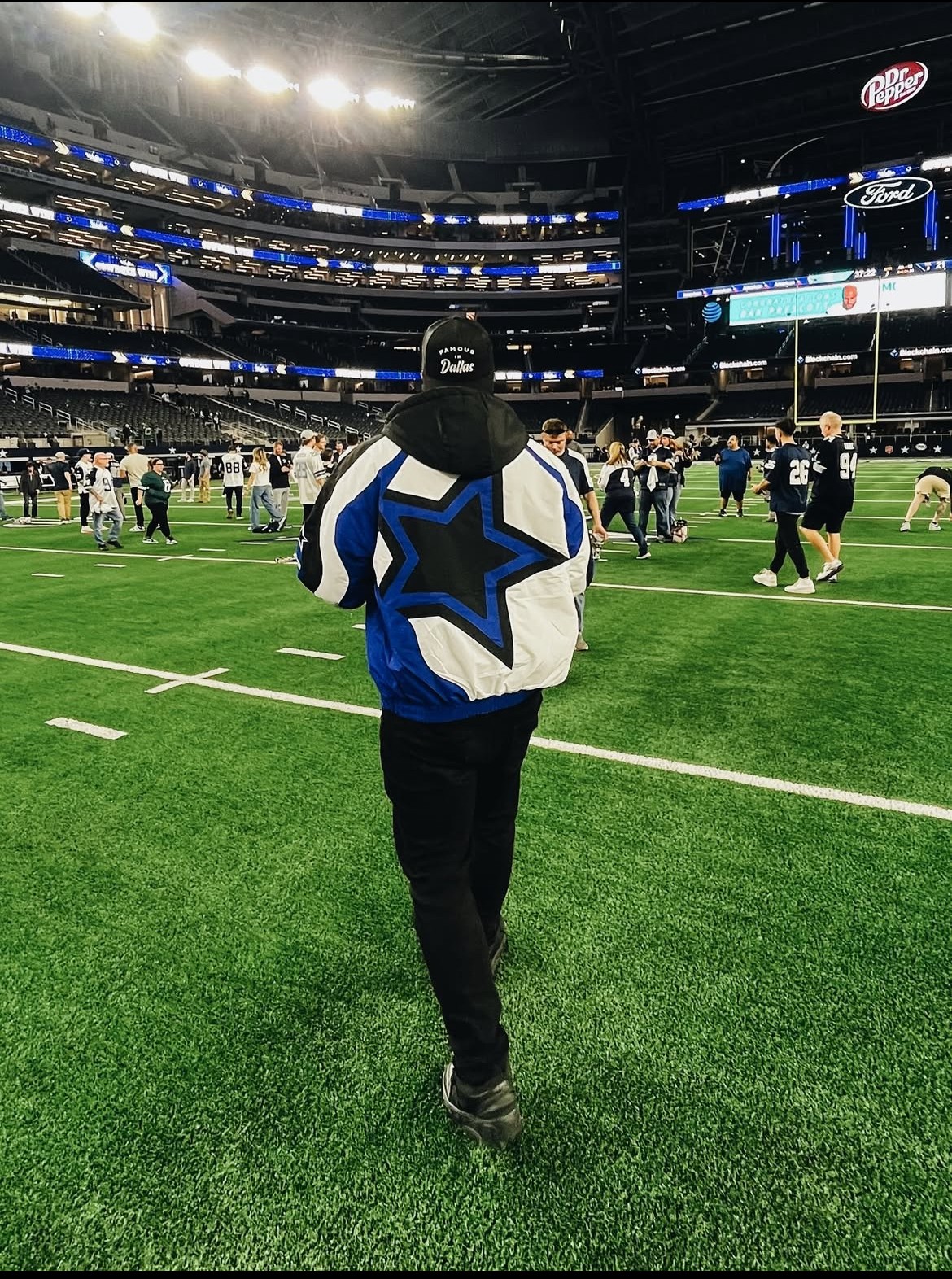 Person walking on a football field wearing a Dallas Cowboys jacket and cap, with a stadium background and people in the field.