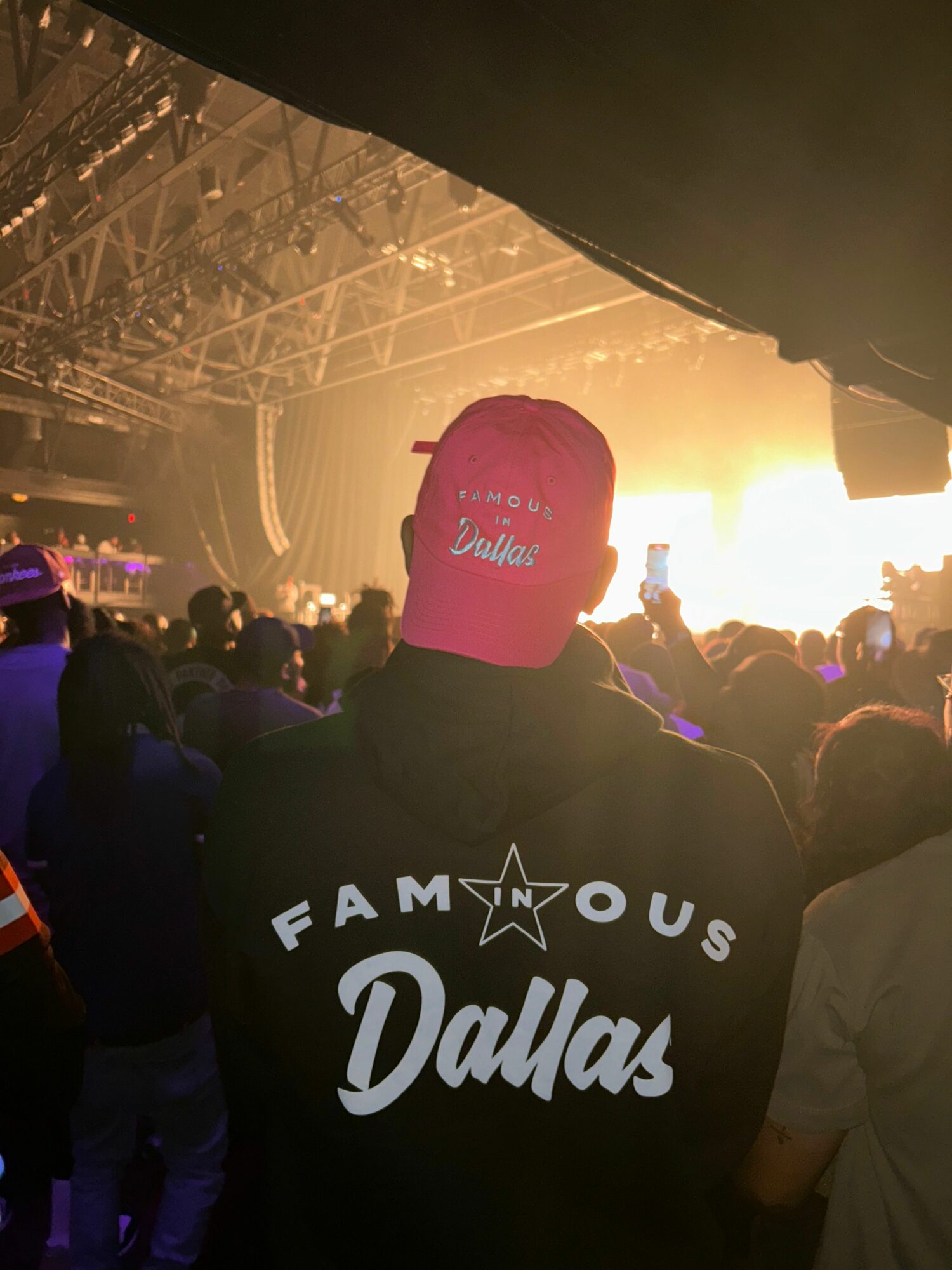 Person wearing a pink cap and black hoodie at a concert with a sunset background, crowd in the distance.
