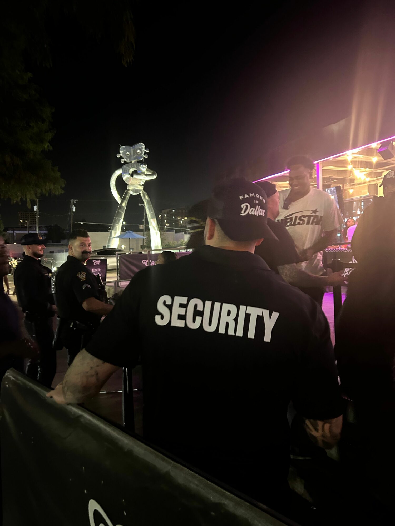 Security guard wearing a black shirt with 'SECURITY' written on the back, standing in a dimly lit area with a trophy and people in the background.